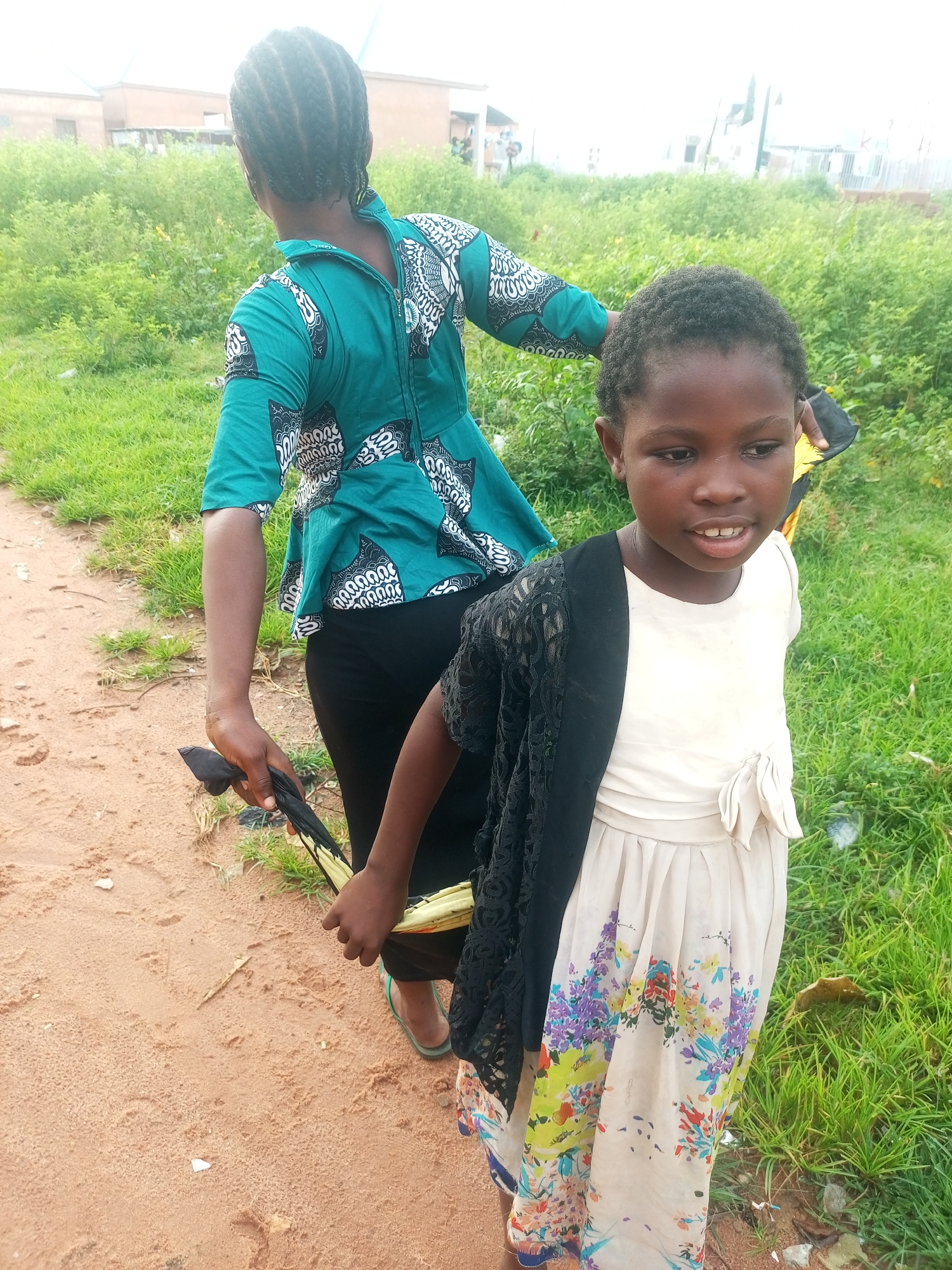 In Tempure, Nigeria, two young Black girls stand in a natural environment. The girl in the foreground, on the right, is wearing a light-colored dress with a floral pattern and a black lace shawl. Her hair is short and textured. The girl in the background, on the left, has her back to the camera and is wearing a green and white patterned top and black pants. Her hair is braided. They are standing on a dirt path next to a grassy area with some vegetation. In the distance, buildings and trees are visible. The lighting suggests it is daytime, possibly overcast.