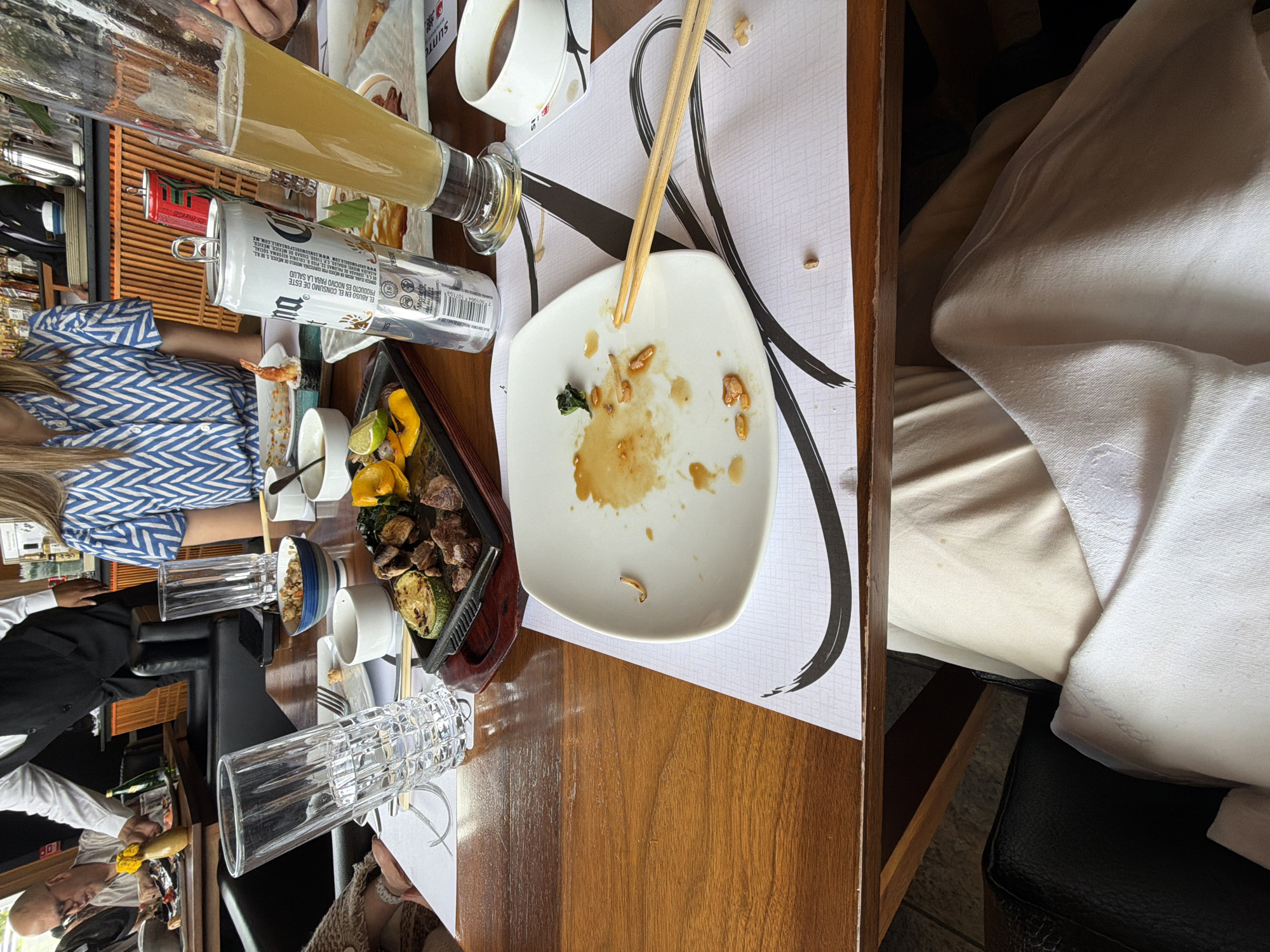 The image depicts a table at a restaurant, possibly in Unknown, Unknown, showing a meal that has been partially consumed. The table is made of wood and is covered with a sheet of paper, presumably a placemat, featuring a design. On the table, there is a square white plate that is mostly empty, containing remnants of food and sauce, along with a pair of chopsticks resting on it. Next to the plate is a cast iron dish filled with grilled food, including meat, zucchini, and yellow peppers, along with small bowls or containers that appear to hold sauces or condiments. A partially consumed can of beer, and another unidentified container, are also visible on the table. To the left, there's a glass of beer and what seems to be the top of a head of a person and their arm, wearing a blue and white shirt. Additional persons are visible at the edge of the frame. The table is set near a padded seat with a white cover, suggesting a comfortable dining environment. The image is taken from an overhead perspective.