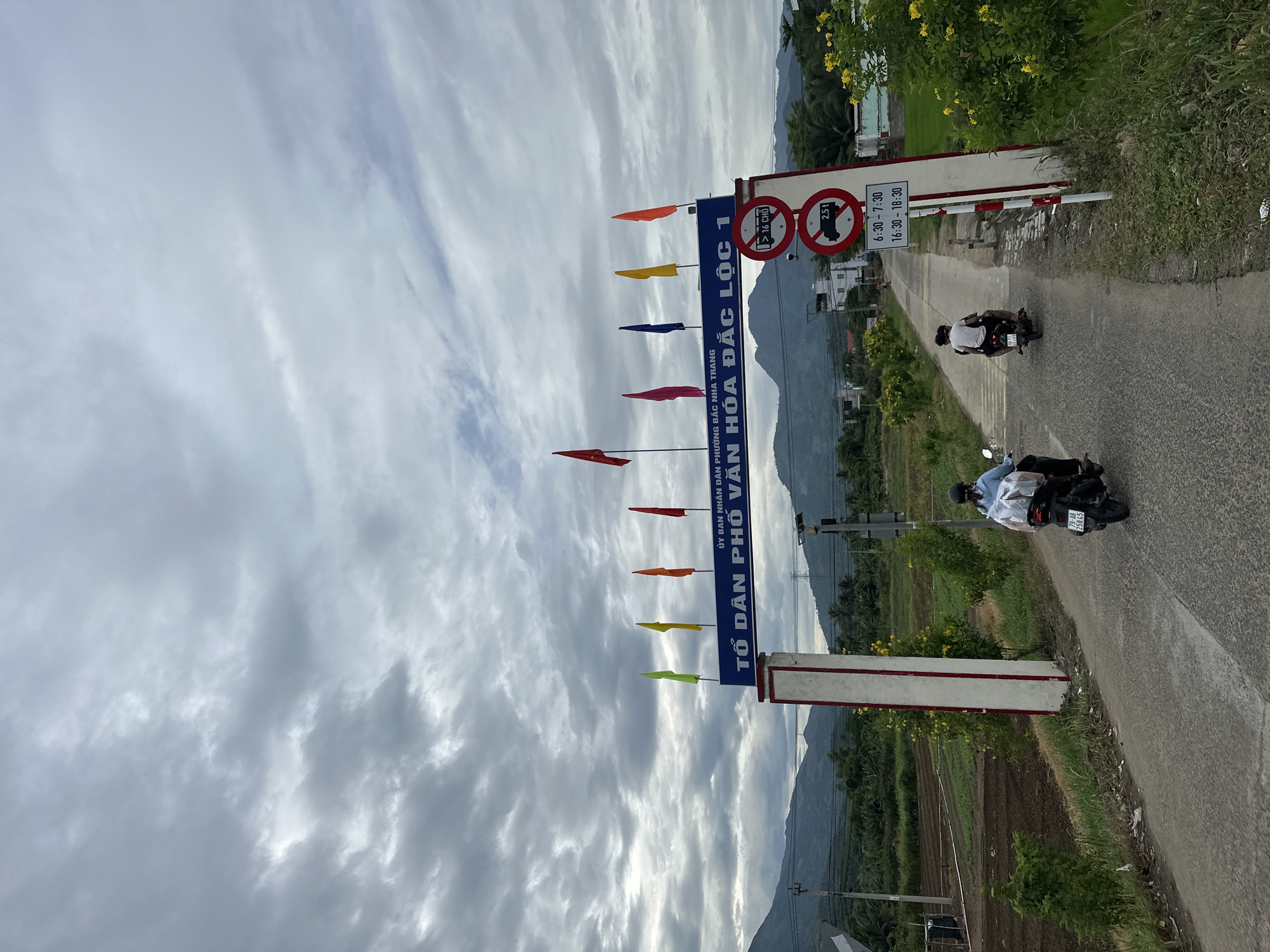 This is a vertical, eye-level, medium shot of a road leading under an archway in a rural area outside of Nha Trang, Vietnam. The archway is adorned with colorful flags and Vietnamese text that reads "ỦY BAN NHÂN DÂN PHƯỜNG BẮC NHA TRANG TỔ DÂN PHỐ VĂN HÓA ĐẮC LỘC 1," translating to "People's Committee of Bac Nha Trang Ward, Cultural Hamlet 1 Dac Loc." Two individuals are riding motorcycles down the road, one in the foreground and another further ahead. The motorcycle in the foreground has a license plate that reads "79-18 258.45." The environment is lush and green, with agricultural fields on the left and trees and buildings on the right. In the background, mountains rise under a cloudy sky. There are traffic signs visible on the right side of the archway, indicating restrictions on vehicles over 2.5 tons and specific operating hours. The overall scene depicts a tranquil, everyday moment in a Vietnamese village.