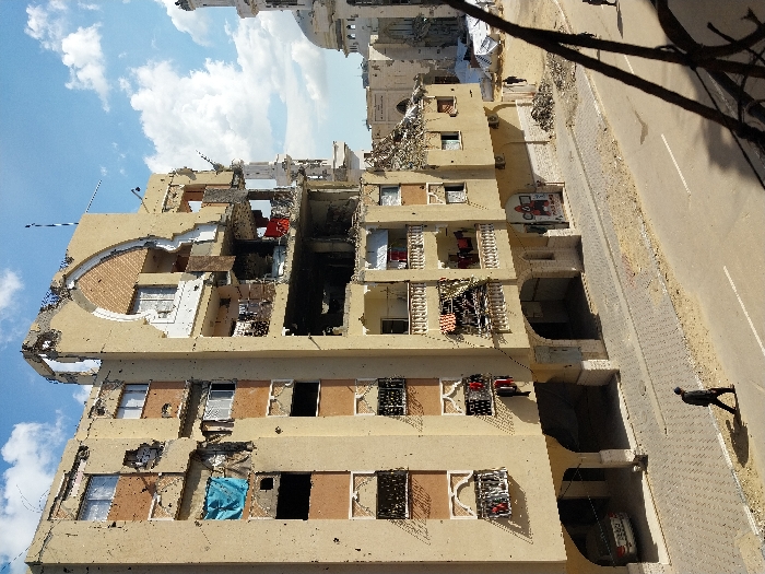 The image depicts a street scene in Khan Yunis, Palestinian Territory, under a partly cloudy sky. The central subject is a heavily damaged, multi-story building, likely residential, exhibiting severe structural compromise on its upper floors and right facade. Large sections of the external walls are missing, exposing interior rooms, and numerous pockmarks consistent with projectile impacts are visible across its tan-colored exterior. Despite the damage, laundry, including a prominent blue item, hangs from balconies and window openings on multiple floors. A white sedan is parked in a recessed, arched ground-floor area of the building.
To the right of this primary building, in the middle ground, stands the Great Mosque of Khan Yunis, identifiable by its white dome and tall, slender minaret. Adjacent to the mosque, further damaged structures and white tarp-covered areas are visible.
At street level, a paved road with a white dividing line runs horizontally across the lower frame, bordered by a tiled sidewalk. One man, dressed in dark clothing, walks from left to right on the sidewalk in the foreground. Another individual is discernible walking further down the street on the right, away from the viewer. A third distant figure is also present near the damaged structures in the background. The immediate bottom-right foreground is partially obscured by out-of-focus dark, skeletal branches or structural elements.