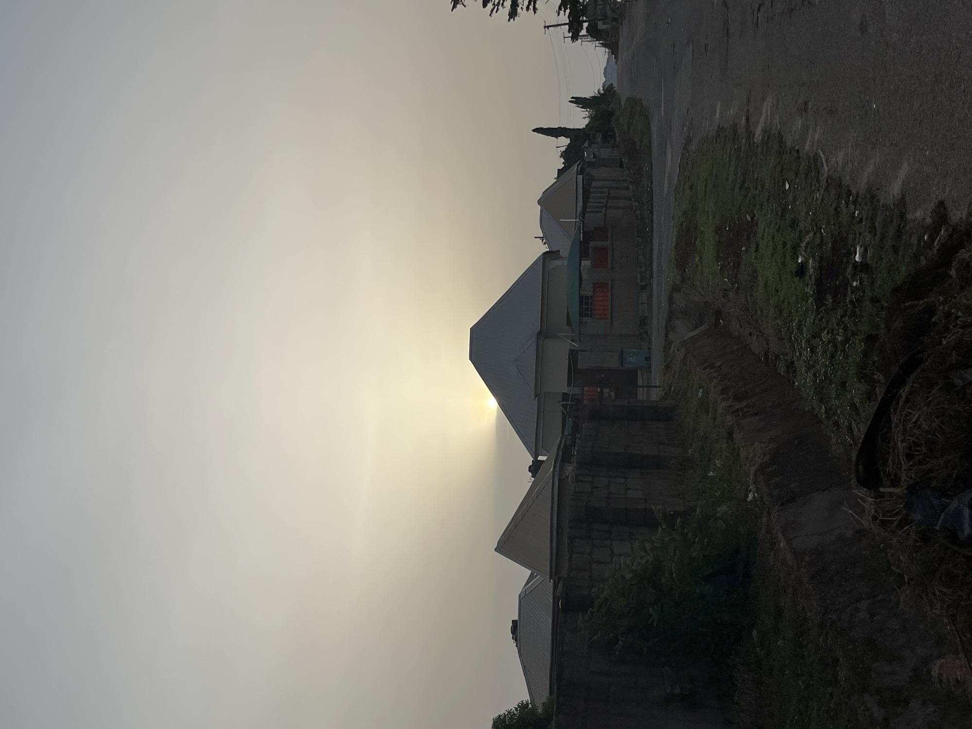 The image depicts a residential area in Abuja, Nigeria, during a time of day when the sun is low in the sky. The light is diffused, creating a hazy or smoky atmosphere. Several houses are visible along a street, with varying roof styles and construction materials. The foreground shows a rough roadside edge with vegetation. The sun is partially obscured, possibly behind a building. There are no people visible in the frame.