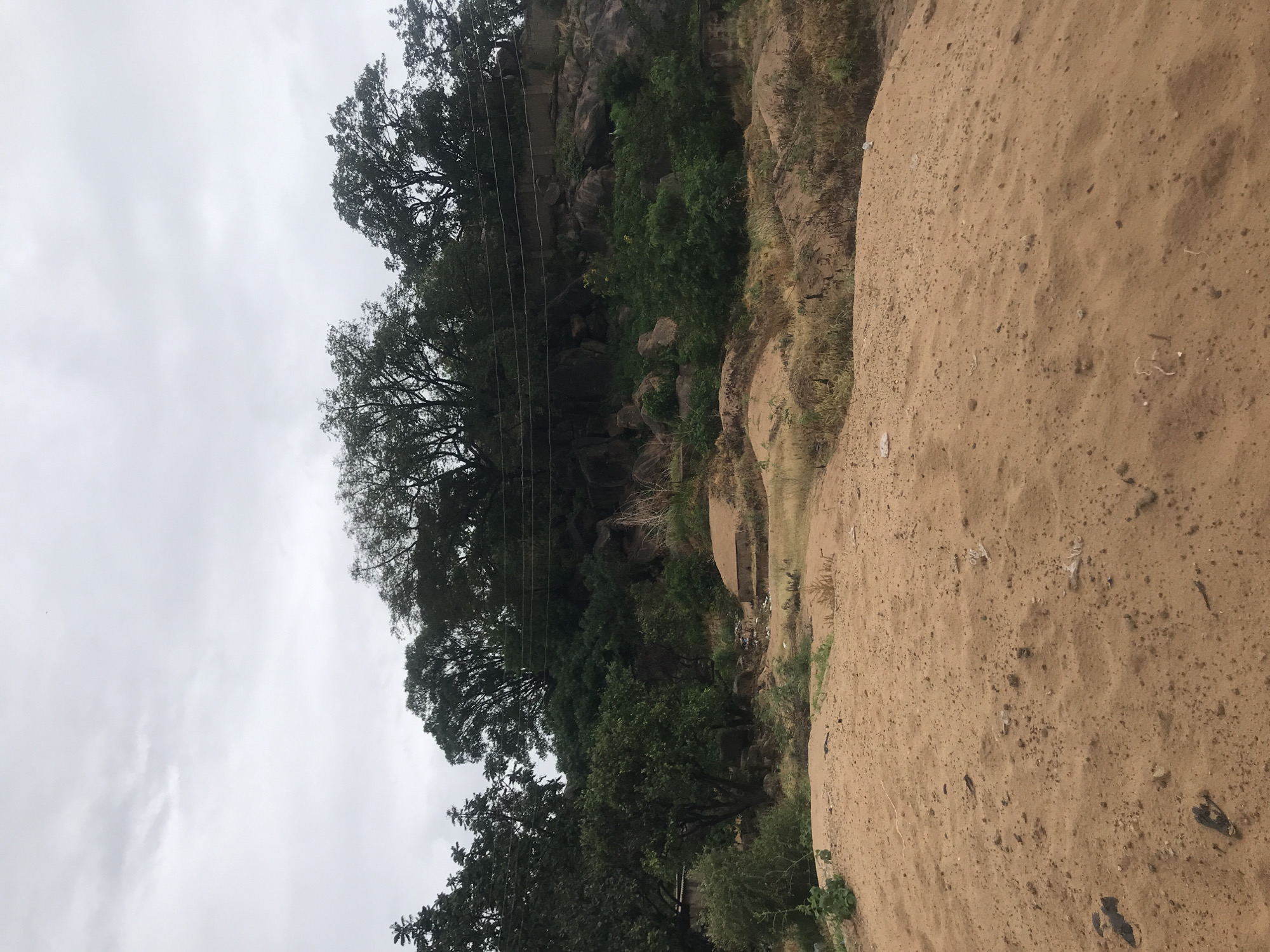 The image shows a dusty, sandy foreground with scattered debris, leading up to a rocky hillside covered in lush green vegetation and trees. Power lines are visible strung across the top of the hill, partially obscured by the foliage. A small, dilapidated concrete structure is visible on the right side of the hill. The sky is overcast with gray clouds. This scene is located in Kabong, Nigeria.