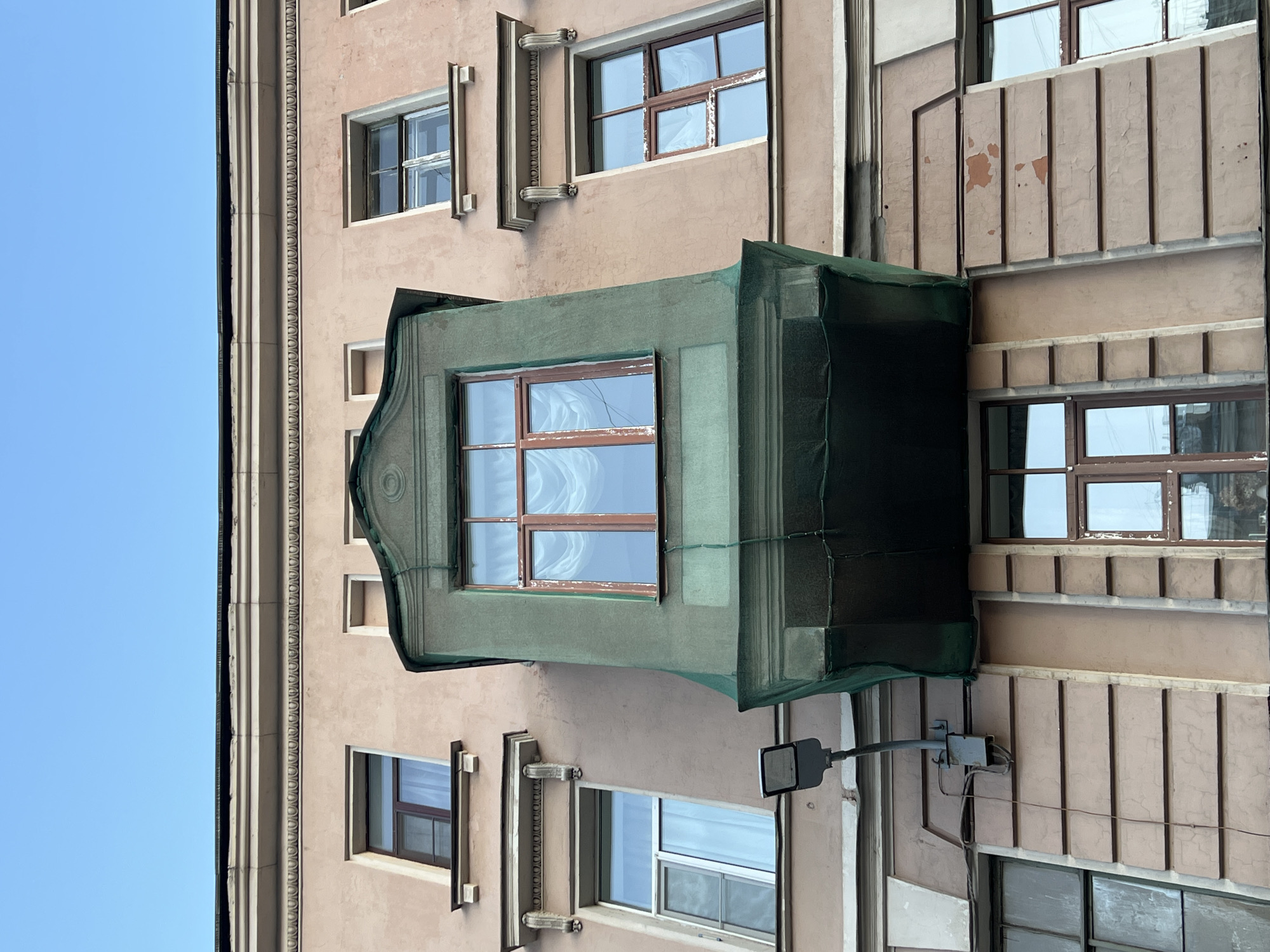 The image depicts a building facade in Saint Petersburg, Russia. The building has a light brown or beige color with architectural details such as decorative moldings and window frames. Several windows are visible, some covered with a green mesh material, possibly for renovation or safety reasons. A street light is mounted on the building's side, with visible wires. The sky is blue and clear. The building appears to be in an urban environment.