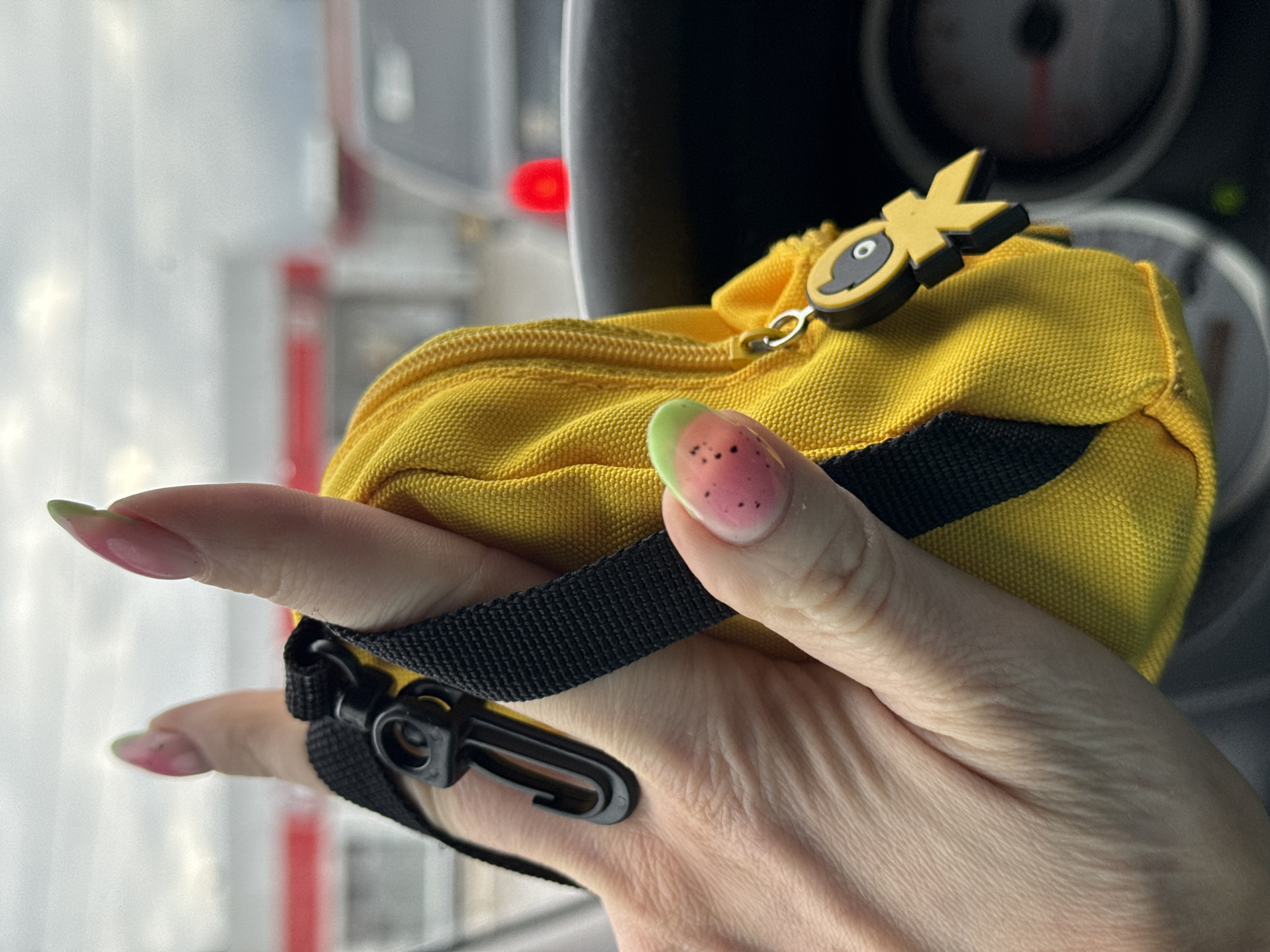 A close-up shot captures a fair-skinned hand holding a small, textured yellow fabric pouch. The hand exhibits long, manicured nails, most featuring an ombre design transitioning from pink to pale green tips. The thumbnail has a distinctive design, showing a pink base with black speckles and a green tip, reminiscent of a watermelon.
The pouch itself is made of a woven material, possibly canvas or nylon, and is bright yellow. A black strap is attached to the pouch, ending in a black plastic carabiner-style hook. A prominent detail on the zipper is a unique pull shaped like the letters "OK," colored yellow and black.
The background is significantly out of focus, indicating a shallow depth of field. Dark, blurry shapes suggest an interior environment, possibly a vehicle cabin, given the appearance of a circular dashboard gauge with a red needle in the upper right, and a blurry red light further back. To the left, a brightly lit, indistinct white and grey area with faint red highlights suggests a view through a window to an outdoor or distant setting.
No specific activity beyond holding the pouch is discernible. The time of day and weather conditions are not clear from the image. The general impression is of a casual moment, possibly taken during travel. The location context provided for this image is Chaikovsky, Russia.