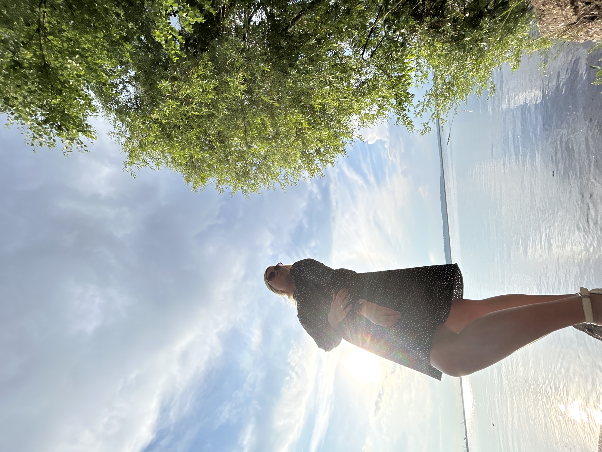 The image depicts a woman standing on a rocky bank near a body of water, likely the Kama River, in Chaikovsky, Russia. She is wearing a dark, polka-dotted dress and sunglasses, and her hair is blonde. The sun is setting, casting a bright glow and creating lens flares. To the right of the frame, a large, green tree with lush leaves hangs over the water. The sky is partly cloudy, with wispy clouds illuminated by the setting sun. The water is calm and reflects the sky and the surrounding landscape. In the distance, a faint shoreline with some industrial structures is visible.