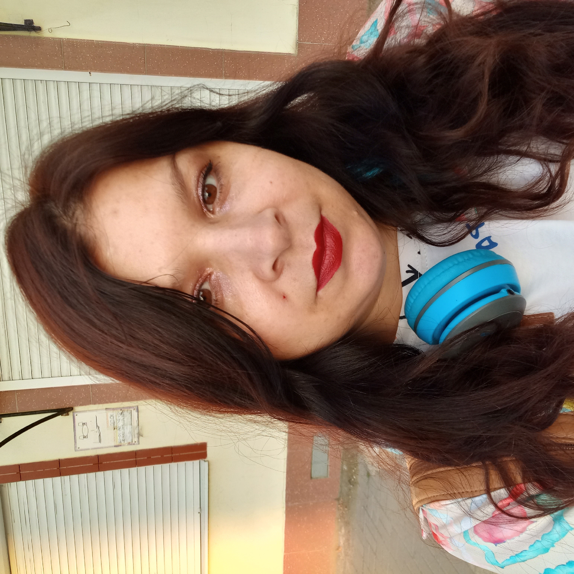 In Kropyvnytskyi, Ukraine, a woman with long, dark brown hair and reddish-brown eyes is captured in a close-up, outdoor selfie. She is wearing a white t-shirt with a colorful abstract pattern and a brown backpack strap is visible over her shoulder. A pair of bright blue headphones rests around her neck. Her lips are adorned with vibrant red lipstick. The background features a building with white roll-up shutters and textured, speckled brown and yellow panels. A sign with a diagram and text is mounted on the wall to the left. The lighting suggests it is daytime, with a warm glow illuminating the scene.