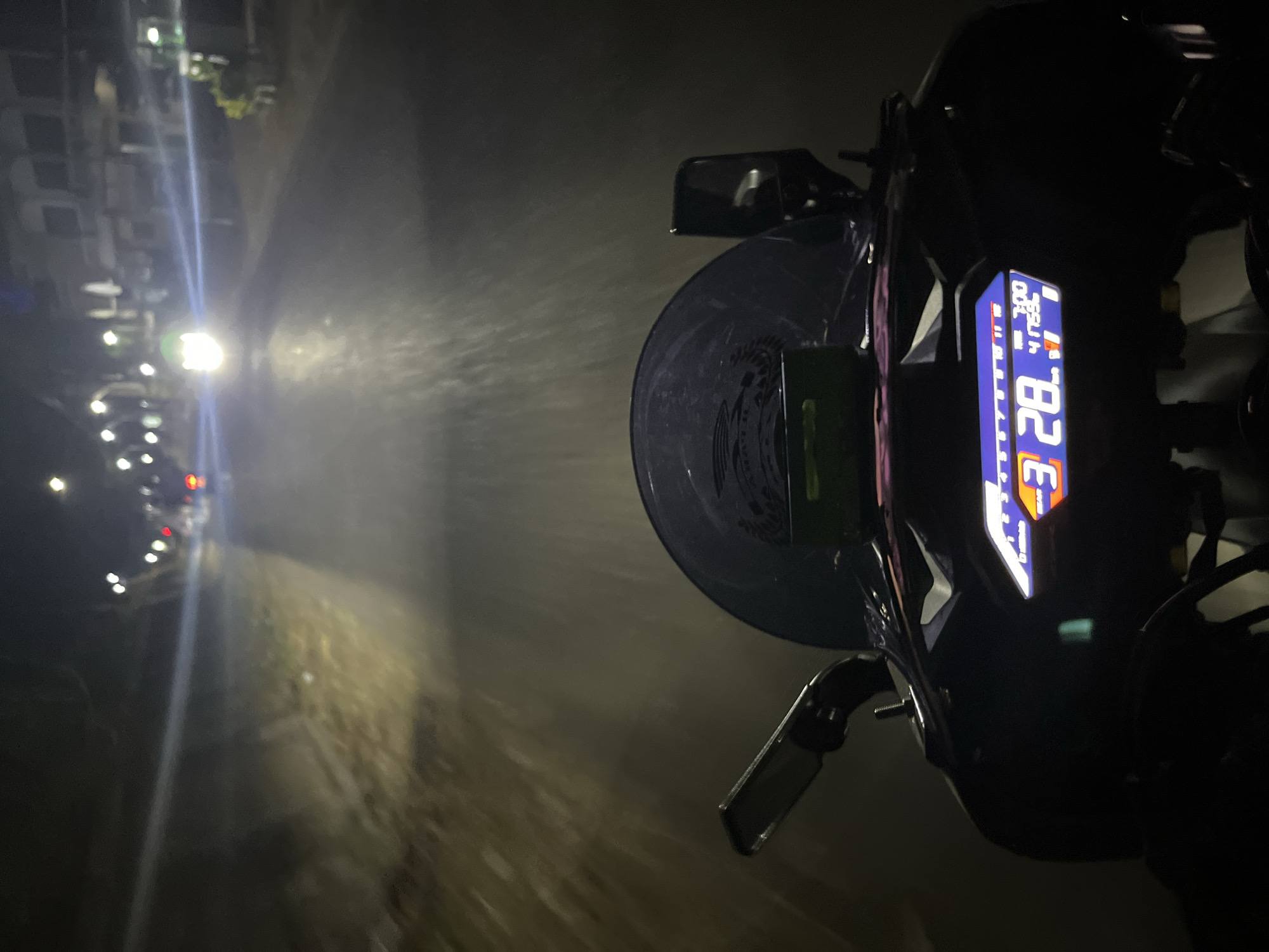 The image depicts a first-person perspective from a motorcycle at night, traveling on a street in Karangrandu, Indonesia. The motorcycle's dashboard displays a speed of 28 km/h and other digital readouts. The road ahead is illuminated by the motorcycle's headlight, revealing a street with buildings and streetlights on either side. Vegetation is visible along the side of the road. The overall atmosphere is dark, typical of a nighttime urban or semi-urban environment.
