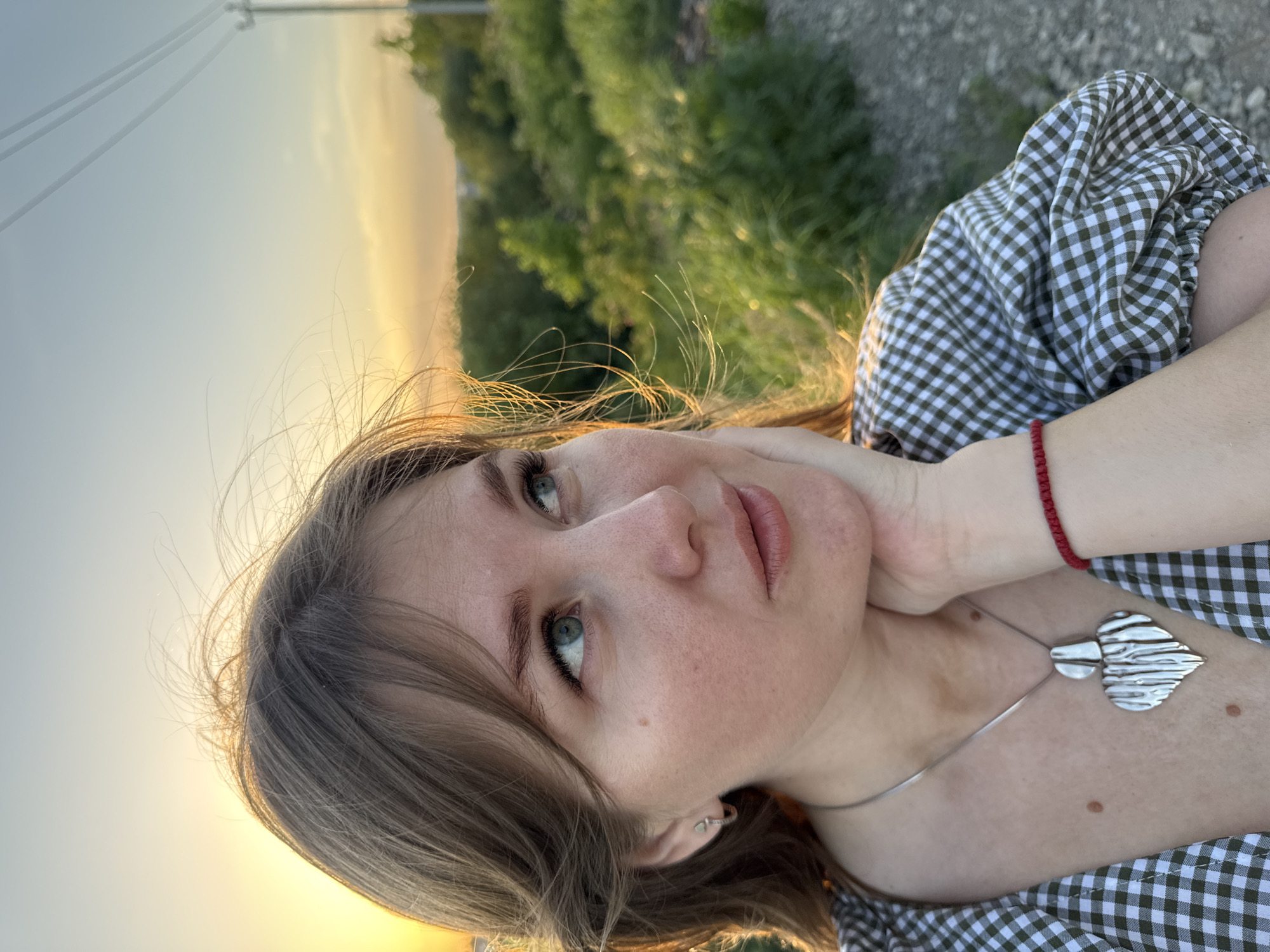 A fair-skinned woman with light brown hair and green eyes is the focus of an outdoor selfie. She wears a green and white checkered blouse, a silver necklace with a distinctive pendant, and a red bracelet. Her left hand rests on her neck. The background is blurred but suggests a natural environment with green foliage and a sky with golden sunlight, possibly near sunset. Overhead power lines are faintly visible in the upper left. The setting is likely in Горный, Russia.