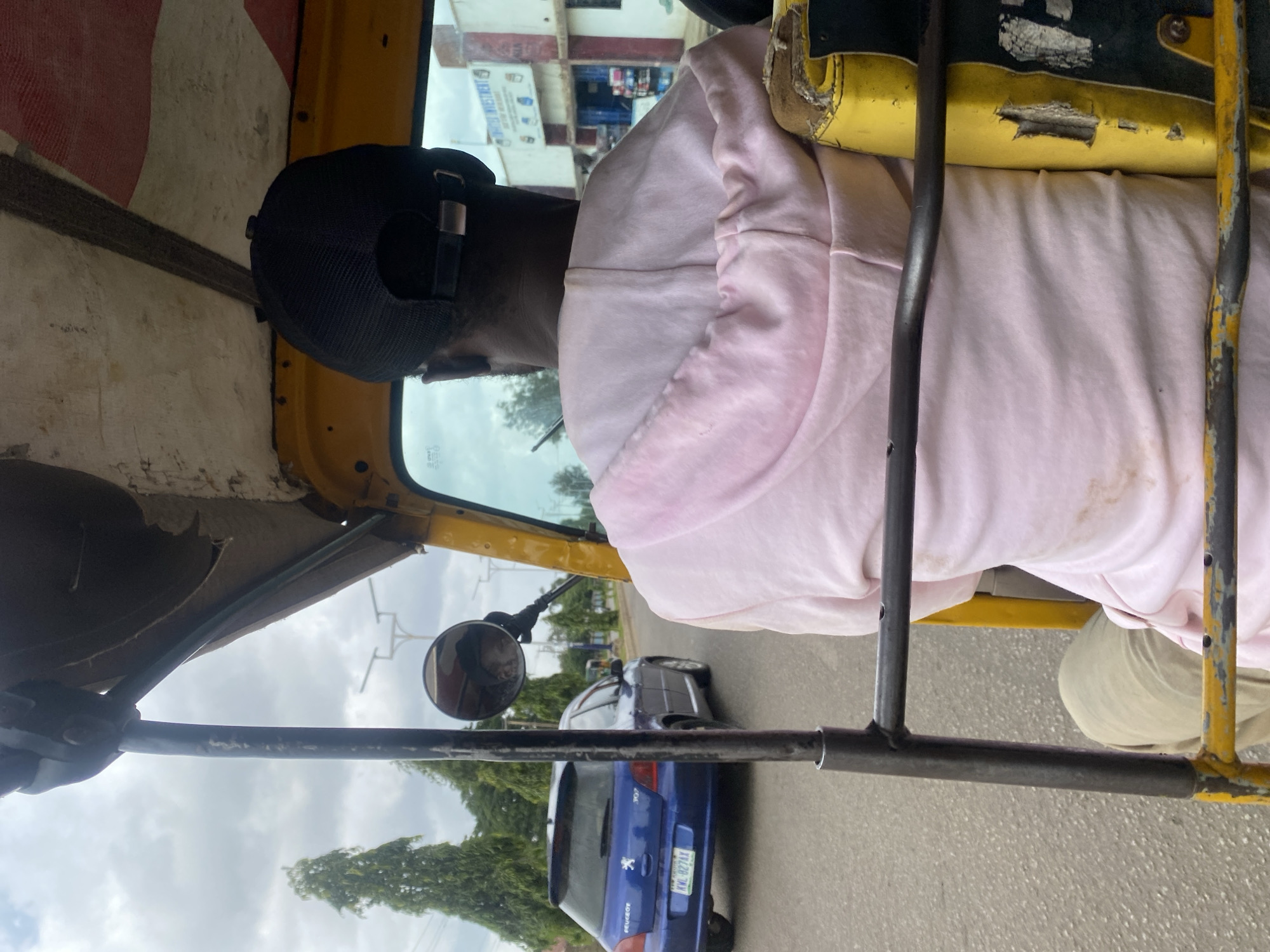 A person wearing a pink hooded sweatshirt and a black baseball cap is seated in the driver's seat of a yellow auto-rickshaw, facing away from the camera. The auto-rickshaw appears to be in motion on a paved road. To the left of the auto-rickshaw, a blue Peugeot 307 hatchback is visible driving in the same direction. In the background, there are trees, a street light, and buildings, suggesting an urban or semi-urban environment. The scene is consistent with a street view in New Stadium, Nigeria.