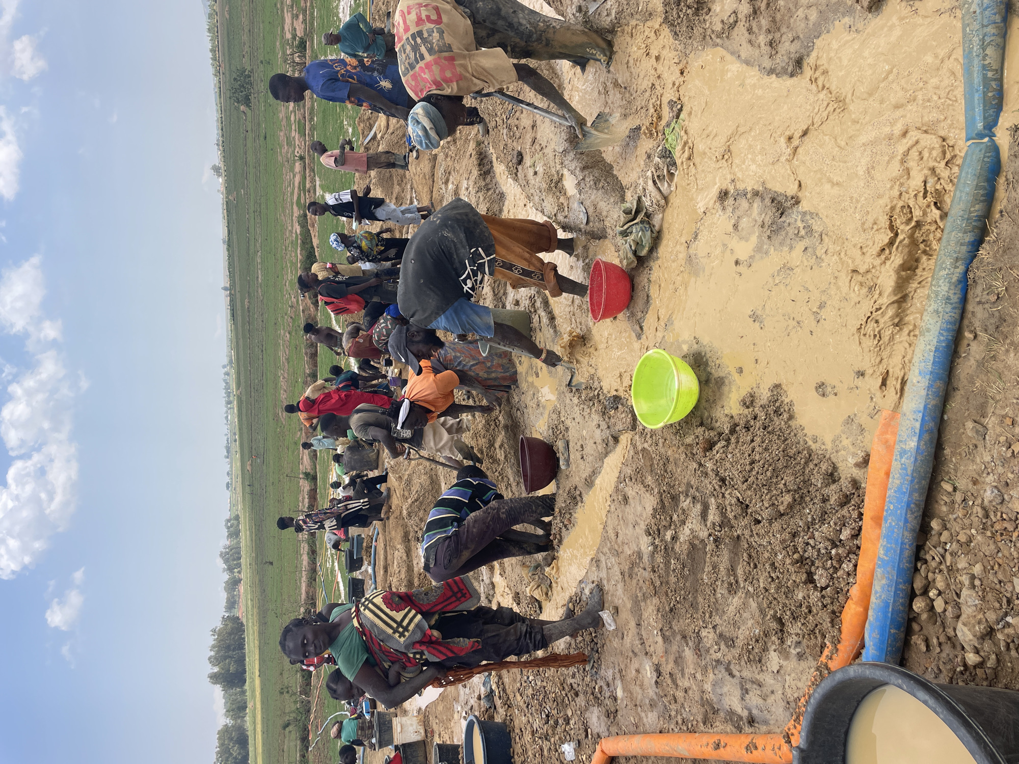 In Maiyanga, Nigeria, a large group of people are digging in a muddy field. They are using shovels and buckets to move the mud, possibly searching for water or other resources. Several blue and orange pipes are visible near the muddy area, suggesting a potential water source or drainage system. The background reveals a flat, green landscape under a cloudy sky. People are positioned in a line within the field, actively involved in the digging process.