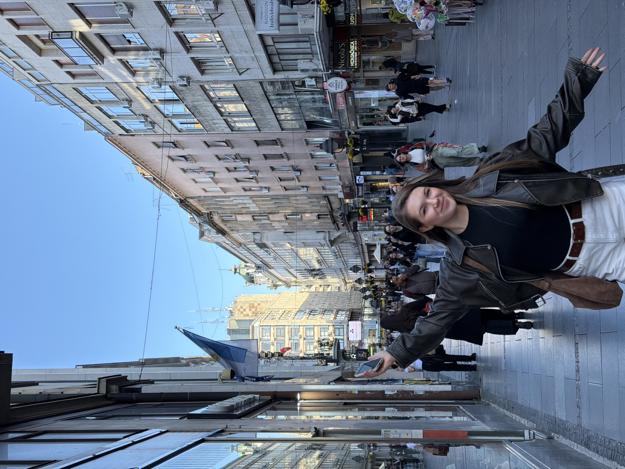 A young woman poses in Knez Mihailova Street, Belgrade, Serbia. She is smiling and extending her arms, wearing a black shirt, brown leather jacket, and white pants. People walk in the background, and shops line the street. The sky is blue, and buildings with multiple windows are visible on either side of the street. The Argentinian flag is seen hanging from a building.