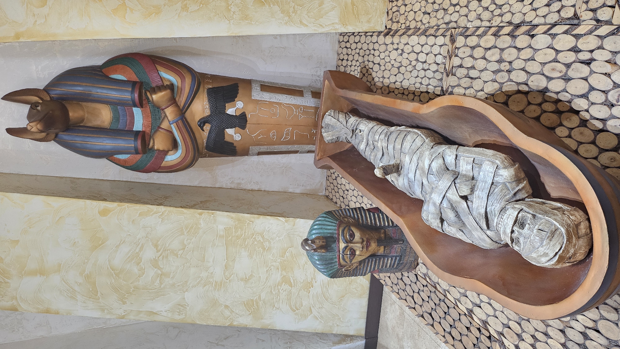 The image depicts an Egyptian-themed display in a room. In the foreground, a mummy is lying in a wooden sarcophagus. To the left of the sarcophagus is a pharaoh's head mask. Behind the sarcophagus, a large statue of Anubis, the jackal-headed god, is standing within a sarcophagus. The floor is covered with a mosaic of circular wooden pieces, and the walls have a textured, yellowish finish. This scene is located in the city of Seogwipo-si, South Korea.