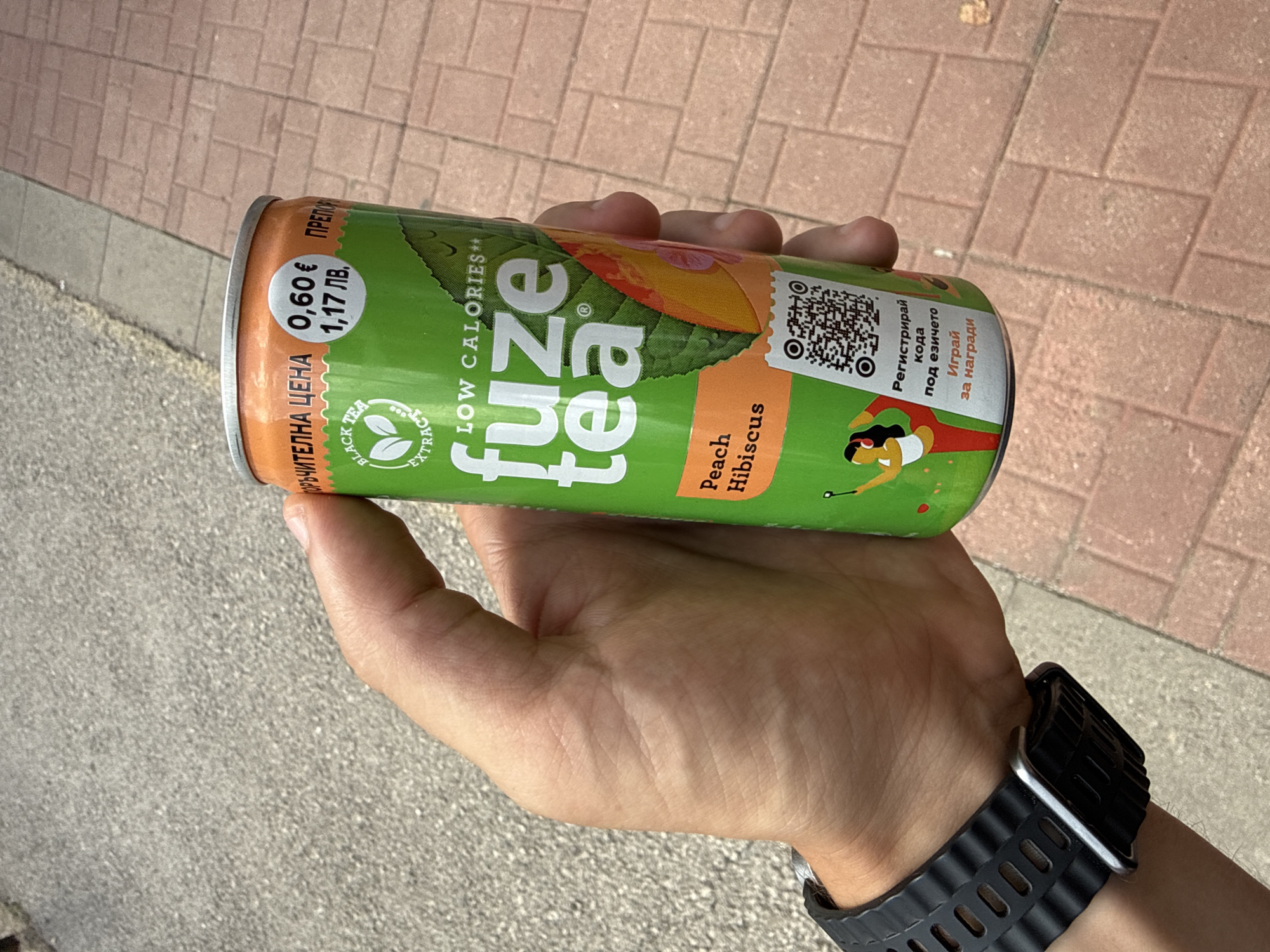 A person's hand holds a green Fuze Tea can outdoors, likely on a street or sidewalk in Vratsa, Bulgaria. The can, prominently featuring the "fuze tea" logo, is a "Peach Hibiscus" flavor and advertises "LOW CALORIES**" and "BLACK TEA EXTRACT." On the left side of the can, price information is visible: "ПРЕПОРЪЧИТЕЛНА ЦЕНА" (Recommended Price) of "0,60 €" (Euros) and "1,17 ЛВ." (Bulgarian Leva). A QR code is present on the right, accompanied by Bulgarian text inviting users to "Регистрирай кода под езичето Играй за награди" (Register the code under the tab Play for prizes). A stylized image of a reclining person is also part of the can's design. The person holding the can wears a black, textured watch with a silver-toned frame on their left wrist. The background shows light-colored paving stones, suggesting a daytime setting with bright, clear weather.
