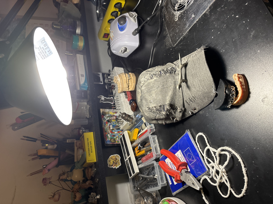 A brightly lit workbench, likely within a workshop or studio in Sofia, Bulgaria, is viewed from an elevated perspective. A black desk lamp, positioned centrally, illuminates the cluttered surface.
In the foreground, a pair of red-handled pliers rests on a blue booklet, encased in plastic, displaying the European Union star logo and the partially visible Bulgarian text "ПОБЕДА - СВЕТОВНО ИЗВЕСТНИ КОЛОДИ ТАРО" (VICTORY - WORLD-FAMOUS TAROT DECKS). A tangled coil of white string lies adjacent to these items. To the right, a dark grey, distressed baseball cap with metal studs and rips is oriented brim-down, with a metallic grey dinosaur figurine positioned on its brim. In front of the cap are two small dental-related objects: a black, irregularly shaped piece and a golden chain-link dental grill resting on a peach-colored gum-line dental model.
Further back on the work surface, a larger tan dental model with wired teeth is visible. To its right, a grey and white electric rotary tool with a blue dial and a connected power cord sits next to a yellow power strip. Various clear plastic organizers contain an assortment of small tools, brushes, tubes of glue or paint, and pens. A circular metal ventilation grill is integrated into the black work surface on the far right.
The background features two shelves. The upper shelf holds dried flowers in a vase, small figurines, a black mesh pen holder containing pencils and brushes, and a prominent yellow box also labeled in Bulgarian "СВЕТОВНО ИЗВЕСТНИ КОЛОДИ ТАРО" (WORLD-FAMOUS TAROT DECKS). This shelf also contains an ink bottle, a green jar, and various other containers. The lower shelf displays a dog sticker, a colorful mosaic-like art piece, and a stand holding numerous small drill bits or burrs. The desk lamp itself bears a label indicating "DESK LAMP Model G28-F3.20 220V-240V~50Hz E27 MAX 60W." No individuals are present.