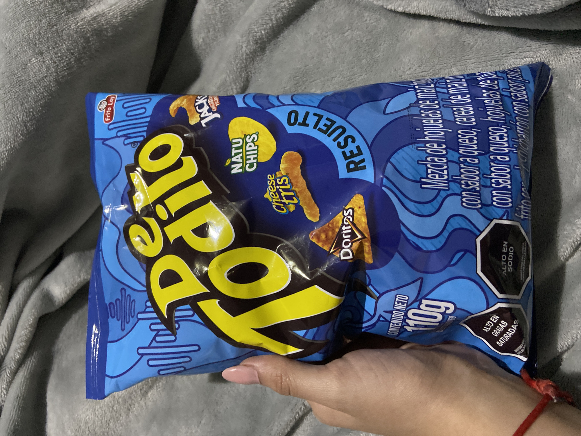 The image displays a close-up of a human hand holding a large, blue Frito Lay snack bag, resting on a soft, grey blanket or textile surface. The hand has light skin, manicured pink nails, and a red string tied around the wrist.
The snack bag features a vibrant blue background with light blue wavy and abstract patterns. The main brand name, "Todito," is prominently displayed in large, yellow, stylized lettering. Above it, the word "RESUELTO" (solved) appears in white, inverted text. Various individual snack brands included in the mix are represented by their logos: "Natu Chips," "Cheese tris," "Doritos," and "Jack's." The Frito Lay logo is visible in the top left corner of the bag.
Text on the bag indicates its contents as a "Mezcla de hojuelas de maiz con sabor a queso, hojuelas de... con sabor a queso, ... chicharrón con sabor" (Mix of corn flakes with cheese flavor, flakes of... with cheese flavor, ... pork rinds with flavor). The net weight is stated as "CONTENIDO NETO 110g." In the bottom right corner, two black octagon warning labels are visible: "ALTO EN SODIO" (high in sodium) and "ALTO EN GRASAS SATURADAS" (high in saturated fats).
The scene suggests a relaxed, indoor setting, possibly in Juan Griego, Venezuela, where this snack product is available. No specific time of day or emotions are discernible.