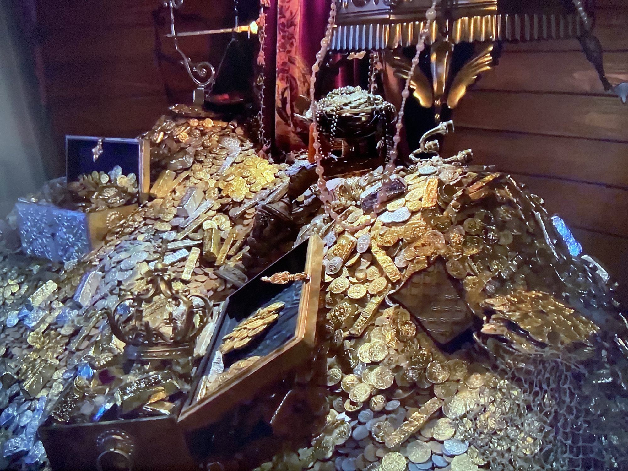 The image depicts a large pile of gold coins and bars, with several ornate chests scattered throughout. The scene appears to be indoors, possibly in a vault or treasure room, based on the wooden paneling visible in the background and the opulent items present. The location is Unknown, Unknown. Dominating the view is a massive mound of golden coins and rectangular bars, suggesting a substantial accumulation of wealth. Several ornate chests, some open and revealing more gold, are positioned within and around the pile. One chest is open with a golden object inside. The items are arranged on the floor with a dark background.