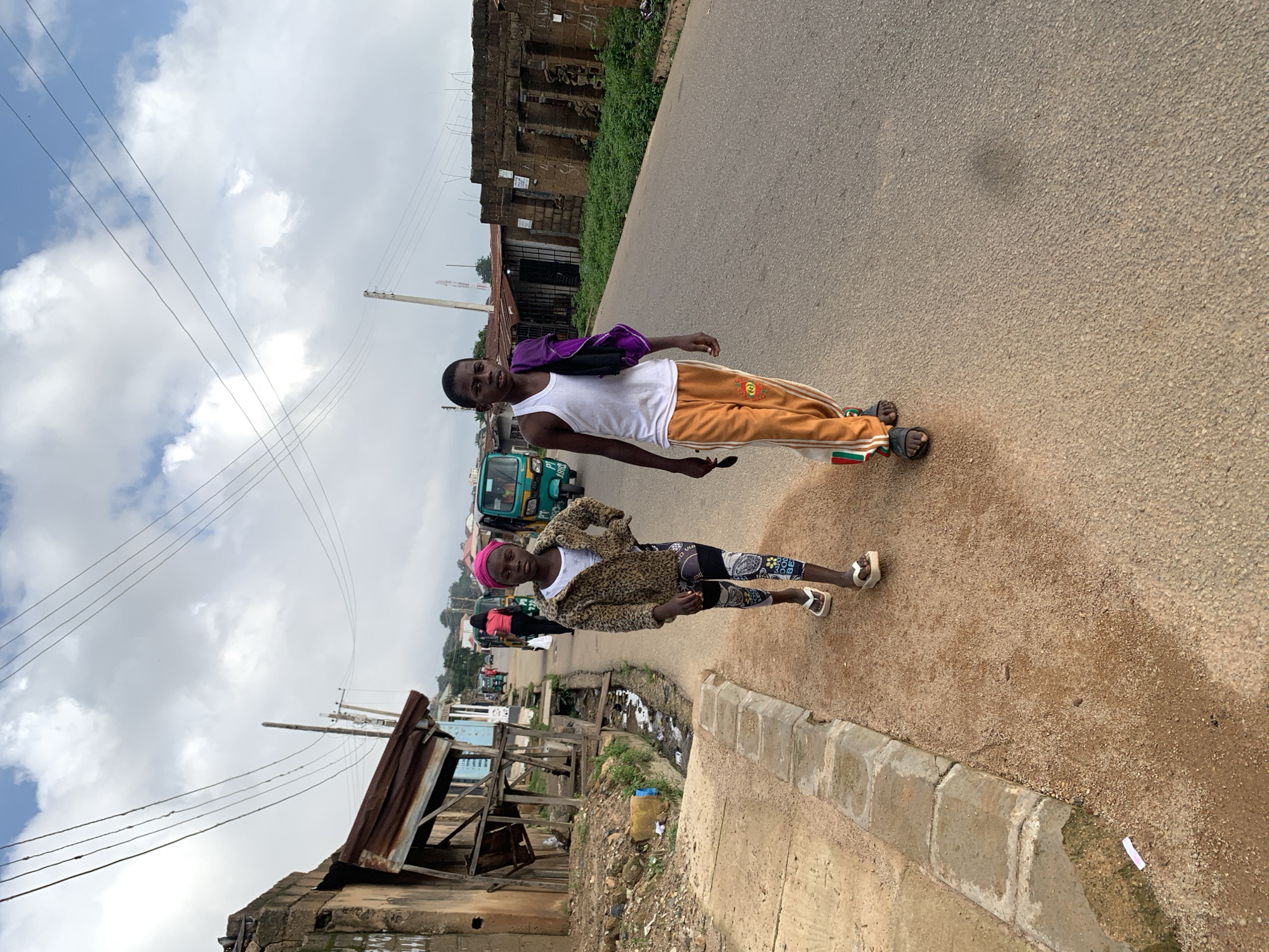 The image shows two children walking on a paved road in Jos, Nigeria. The child on the left, a young girl, wears a leopard-print jacket, a white t-shirt, patterned leggings, and white flip-flops. She also has a pink headscarf. The child on the right, a young boy, wears a white tank top, orange pants with green and red stripes, and dark sandals. He carries a purple cloth draped over his shoulder. They are walking on the right side of the road, facing forward.
The environment appears to be a street with low-rise buildings on both sides. On the left, there are structures with corrugated iron roofs and some vegetation. On the right, there are more weathered buildings, some appearing partially constructed or in disrepair. In the background, several motorized tricycles, commonly used as taxis in Nigeria, are visible, along with utility poles and overhead wires. The sky is partly cloudy, with a mix of blue sky and white clouds. The road surface is asphalt, with a sandy shoulder on the left side. The overall scene suggests a typical urban or semi-urban street in Jos.