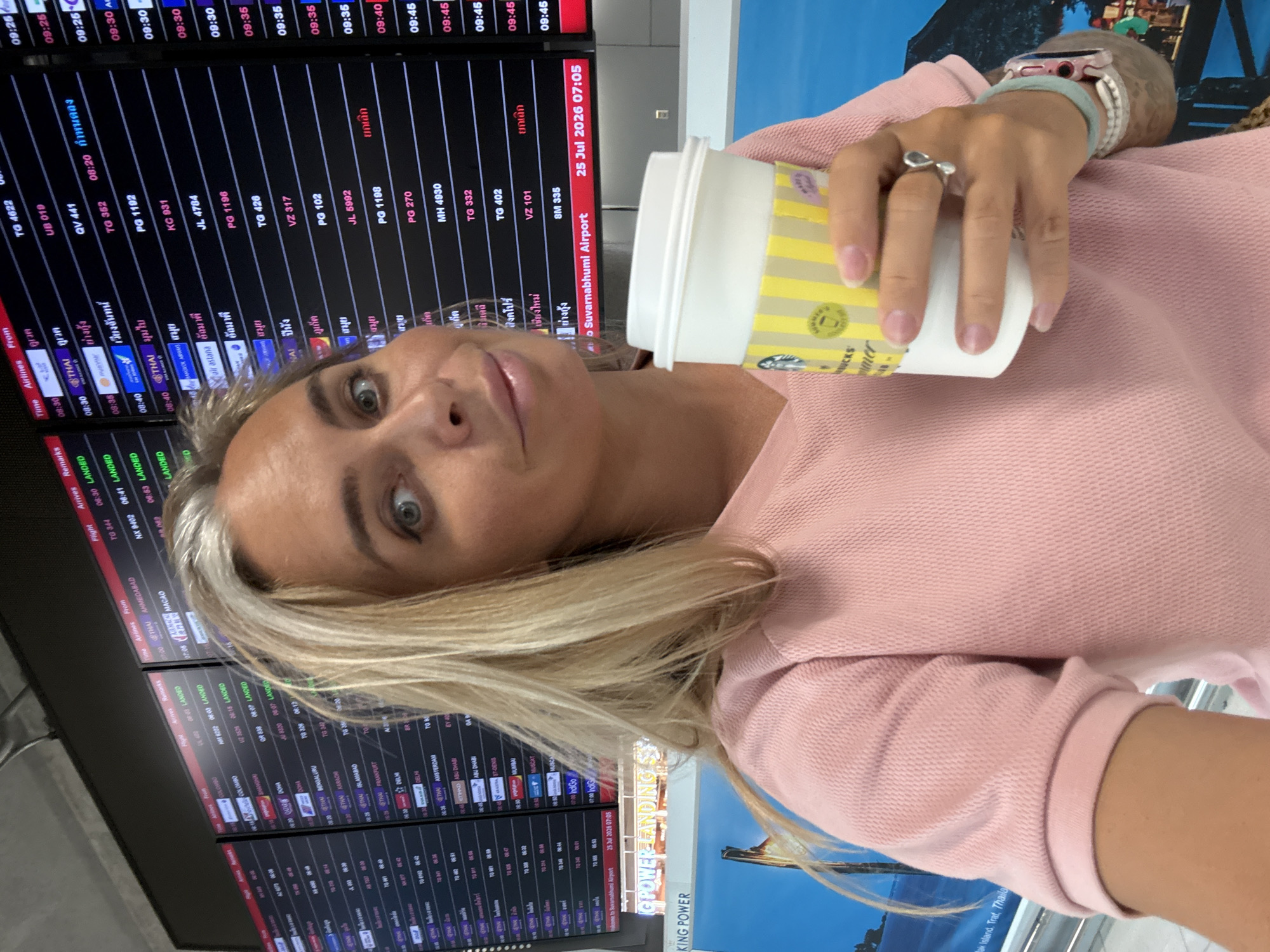A woman with blonde hair holds a Starbucks cup and smiles at the camera. She is wearing a pink shirt. In the background, an airport departure board displays flight information. The departure board indicates that the location is Suvarnabhumi Airport, with a date of July 25, 2026, at 07:05. Suvarnabhumi Airport is located in Bang Phli District, Samut Prakan Province, near Bangkok, Thailand. The city of Nong Prue Subdistrict is not directly indicated by the visual elements in the image.