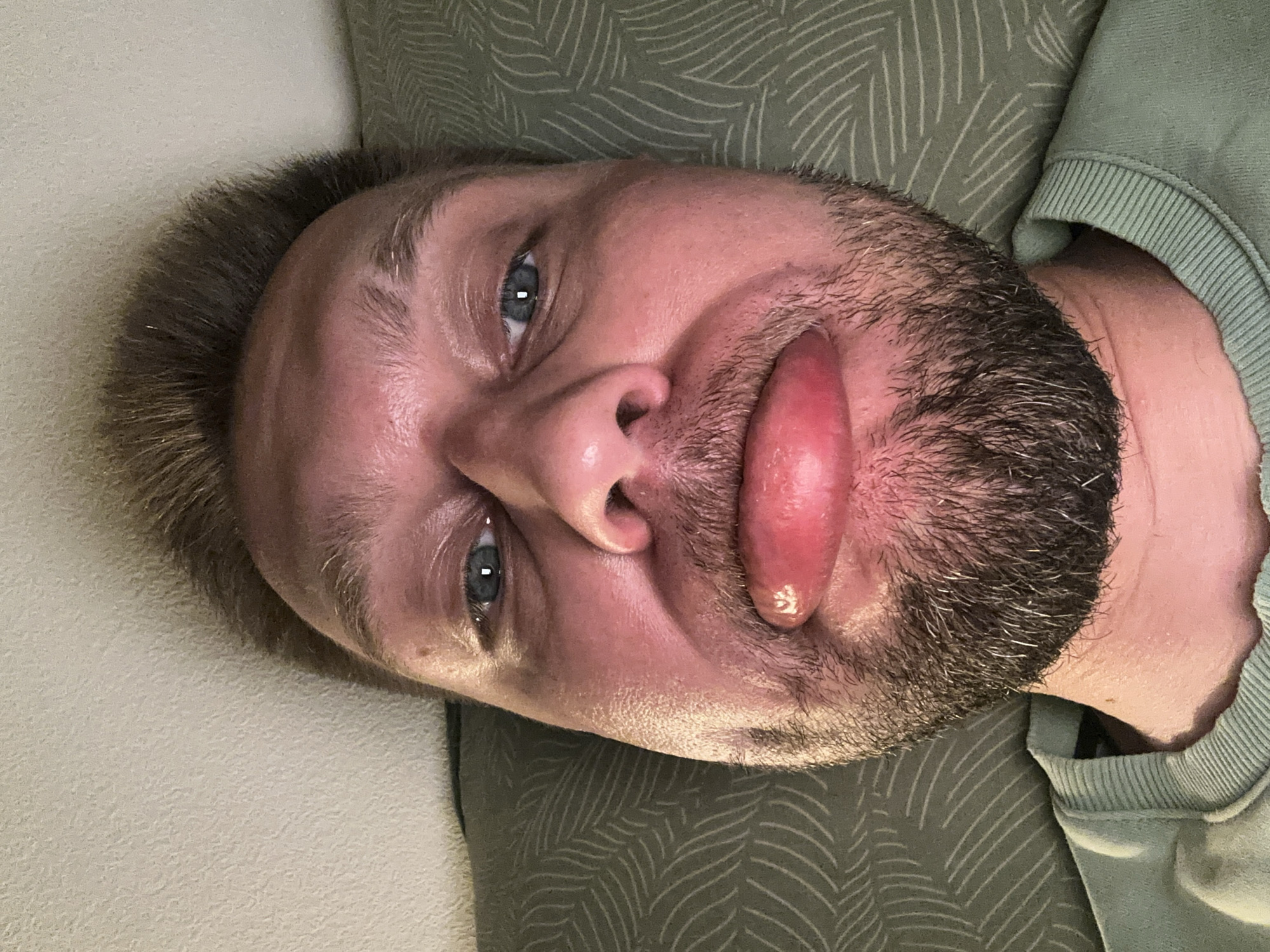 This is a close-up, rotated image of a man's face, likely taken indoors. The man has light skin, blue eyes, and short, light brown hair. He has a beard with darker hairs and some gray streaks. His mouth is open, and his tongue is extended and appears swollen or discolored, with a reddish-pink hue. There is some light visible on his forehead and nose, suggesting it might be daytime.
The man is lying down, propped up by pillows. One pillow is a light, textured cream color, while the other is a dark green fabric with a repeating leaf-like pattern. He is wearing a light green crew-neck sweatshirt. The lighting in the image is soft, creating a warm tone on his skin. The focus is on his face, particularly his eyes and mouth. There is no visible text in the image. The overall impression is a casual, perhaps slightly humorous, self-portrait. The setting appears to be a domestic environment, possibly a bedroom or living room.