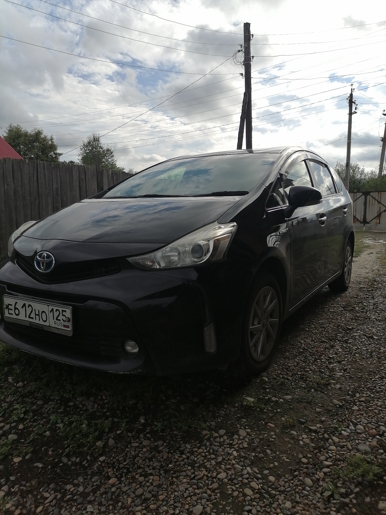 A black Toyota Prius, license plate E612HO125 RUS, is parked on a gravel surface. The car is positioned at a slight angle, facing left and towards the viewer. In the background, a wooden fence and several utility poles with overhead wires are visible. The sky is partly cloudy. The scene appears to be in Novopokrovka, Russia, given the license plate.