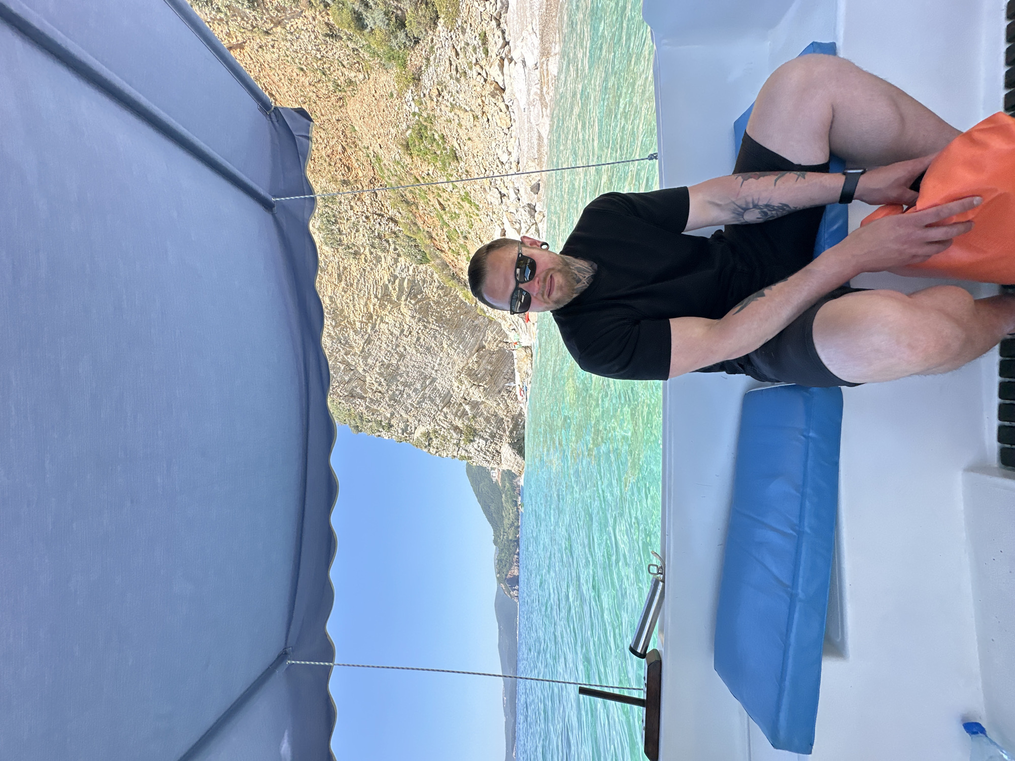 A man with a beard and sunglasses sits on a white boat on the turquoise sea in Budva, Montenegro. He wears a black t-shirt and black shorts, with tattoos visible on his arm. He is holding an orange bag. Behind the boat, a rocky coastline rises from the water. A blue sunshade covers part of the boat. Blue cushions are on the boat's seating area.