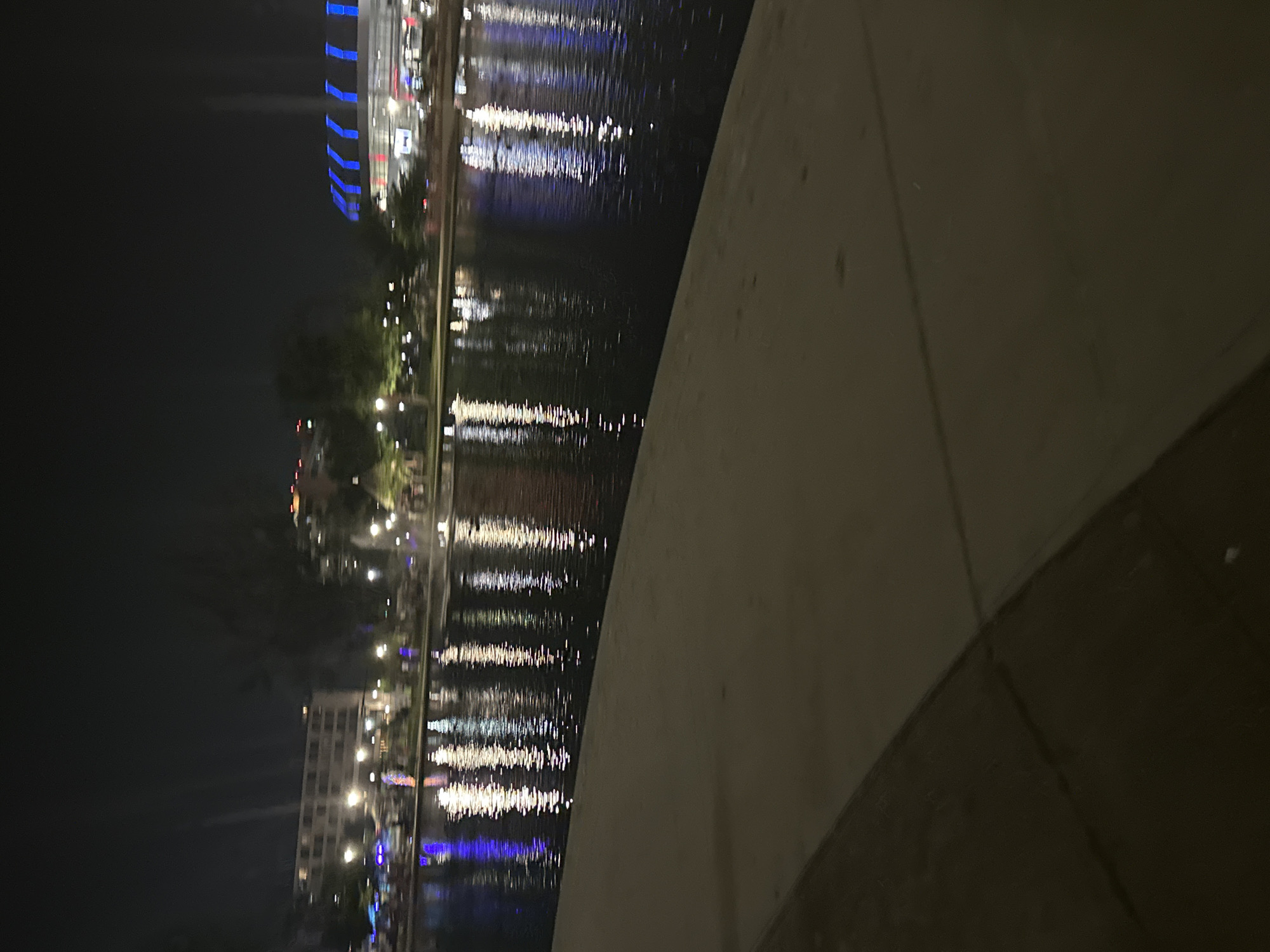 The image depicts a nighttime scene at Bridgestone Park in Huntsville, Alabama. A body of water, reflecting the lights of surrounding buildings and a stadium, occupies the majority of the frame. The water's surface is rippled, distorting the reflections into vertical streaks of white and blue. In the background, the distinct architecture of a stadium with blue and white illuminated features is visible on the right. To the left, a multi-story building with numerous lit windows is apparent, along with streetlights and trees. The foreground consists of a paved walkway with a curved edge, leading down to the water's edge. The overall atmosphere is dark, with artificial lights creating a vibrant contrast against the night sky. No people are clearly discernible in the image.