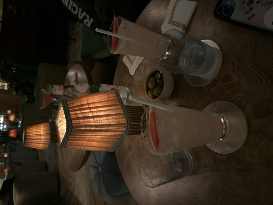 The image captures a dimly lit, cozy indoor setting, likely a bar or lounge, with a focus on a wooden table laden with drinks and snacks. The overall impression is one of relaxation and ambiance.
On the dark, wood-grain patterned table, two tall cocktail glasses are prominent. Each glass contains a light pink or peach-colored beverage, garnished with what appears to be a slice of grapefruit or watermelon, and a white straw. The rims of both glasses are coated with salt or sugar. Both glasses rest on round, possibly metallic or reflective, coasters.
Between the two main drinks, there's a small, white ceramic bowl filled with green olives, some of which appear to have pimentos, with a few picks inserted. Adjacent to the olives is a clear glass bottle of San Pellegrino sparkling water, clearly labeled "SAN PELLEGRINO."
Two decorative table lamps with pleated, ribbed shades are positioned on the table, casting a warm, orange glow. The lamps are illuminating the immediate area around the drinks and the water bottle. A white cloth napkin with a decorative border is also visible near one of the cocktail glasses.
In the upper right corner of the frame, part of a smartphone with a distinctive case (showing a cartoon-like character or design) and camera lenses is visible.
The background shows more of the establishment's interior, featuring plush seating, including a dark green upholstered chair or booth, and a light blue/grey tufted chair. There are blurred lights further in the background, adding to the ambient atmosphere. Part of a person's dark clothing with white, possibly reversed, text (appearing to be "RACING") can be seen behind one of the drinks.
The scene suggests an evening or night out, with individuals enjoying drinks and conversation in a sophisticated and comfortable environment.
