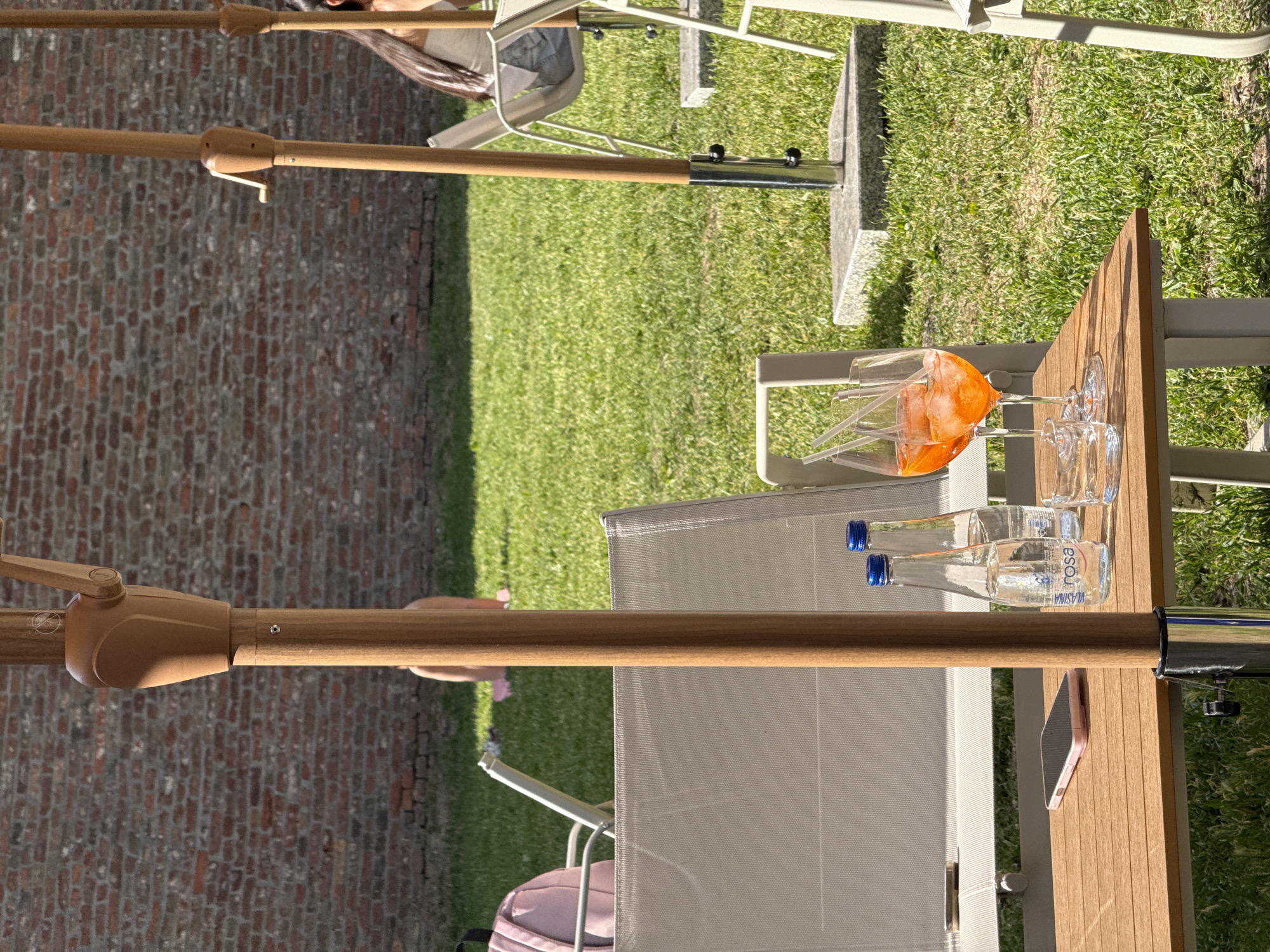 The image captures an outdoor setting, likely in Belgrade, Serbia. A patio table is visible, on which sits a partially consumed orange drink in a glass, along with two small water bottles labeled "VLASINA ROSA", and a cell phone. The table is situated on a grassy area next to a brick wall. The scene seems to be at an establishment (perhaps a restaurant or cafe) with patio furniture set up for patrons. There are chairs and umbrellas visible in the background, with hints of people enjoying the outdoor space. The atmosphere suggests a relaxed summer day.