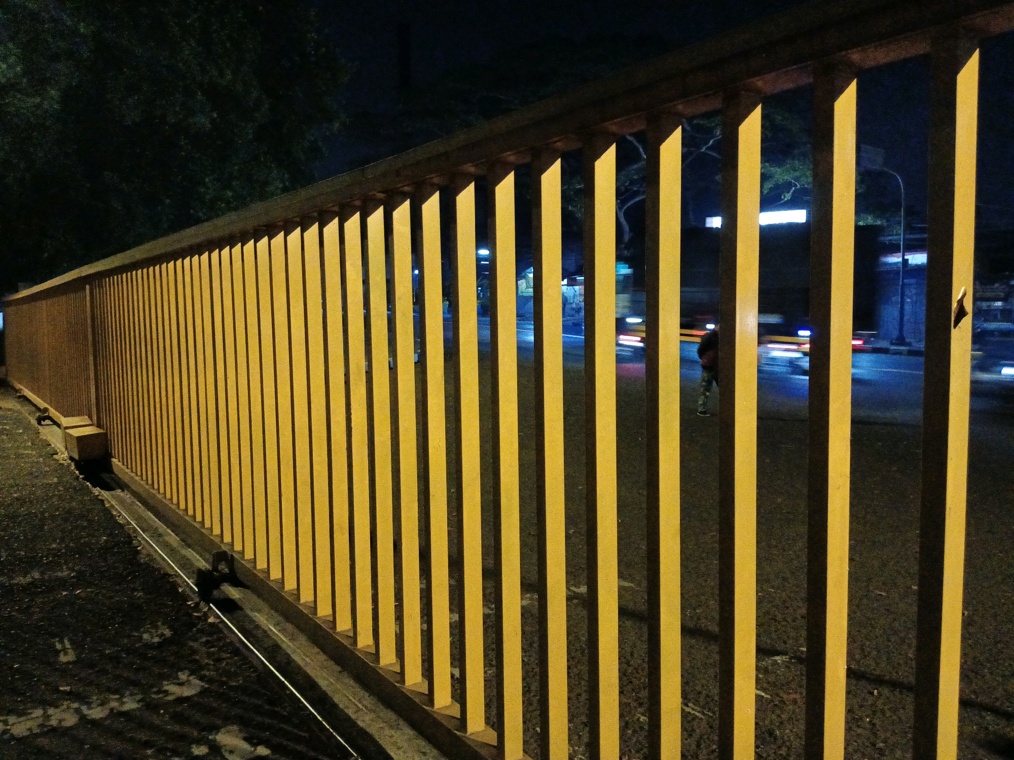 This image captures a nighttime view of a bright yellow metal fence, likely a railing along a sidewalk or bridge. The fence is composed of vertical bars spaced evenly apart, creating a repetitive pattern that dominates the foreground. Beyond the fence, a street is visible with blurred vehicle lights, indicating movement and activity. The background consists of dark trees and some illuminated structures, suggesting an urban environment. The lighting is artificial, casting shadows from the fence bars onto the pavement. There is a single person visible in the distance, partially obscured by the fence, walking on the street. The overall scene suggests a quiet, late-night urban setting in Bandung City, Indonesia, characterized by the stark contrast between the illuminated fence and the dark, active street beyond.