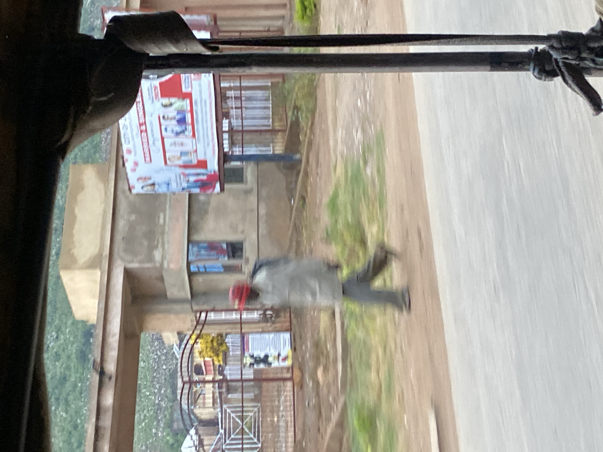 The image depicts a street scene in Gwong, Nigeria, possibly near an entrance to a complex. In the foreground, a portion of a vehicle, likely a tuk-tuk or similar motorized rickshaw, is visible on the right, with dark metal poles and ropes, indicating it is the viewpoint of someone traveling within. A blurred figure of a man wearing a red cap, a light-colored jacket, and dark trousers is walking from left to right across the scene. He appears to be walking on a dirt or gravel path adjacent to a paved road. Behind the man, there is an arched gateway with concrete structures and a fence. A billboard is mounted on a structure to the right of the gateway, displaying text and images, with "CITY OF..." and "MARRIED & SINGLE" visible. The background shows hilly terrain covered with vegetation. The overall scene is slightly out of focus due to motion blur, suggesting the image was taken from a moving vehicle.