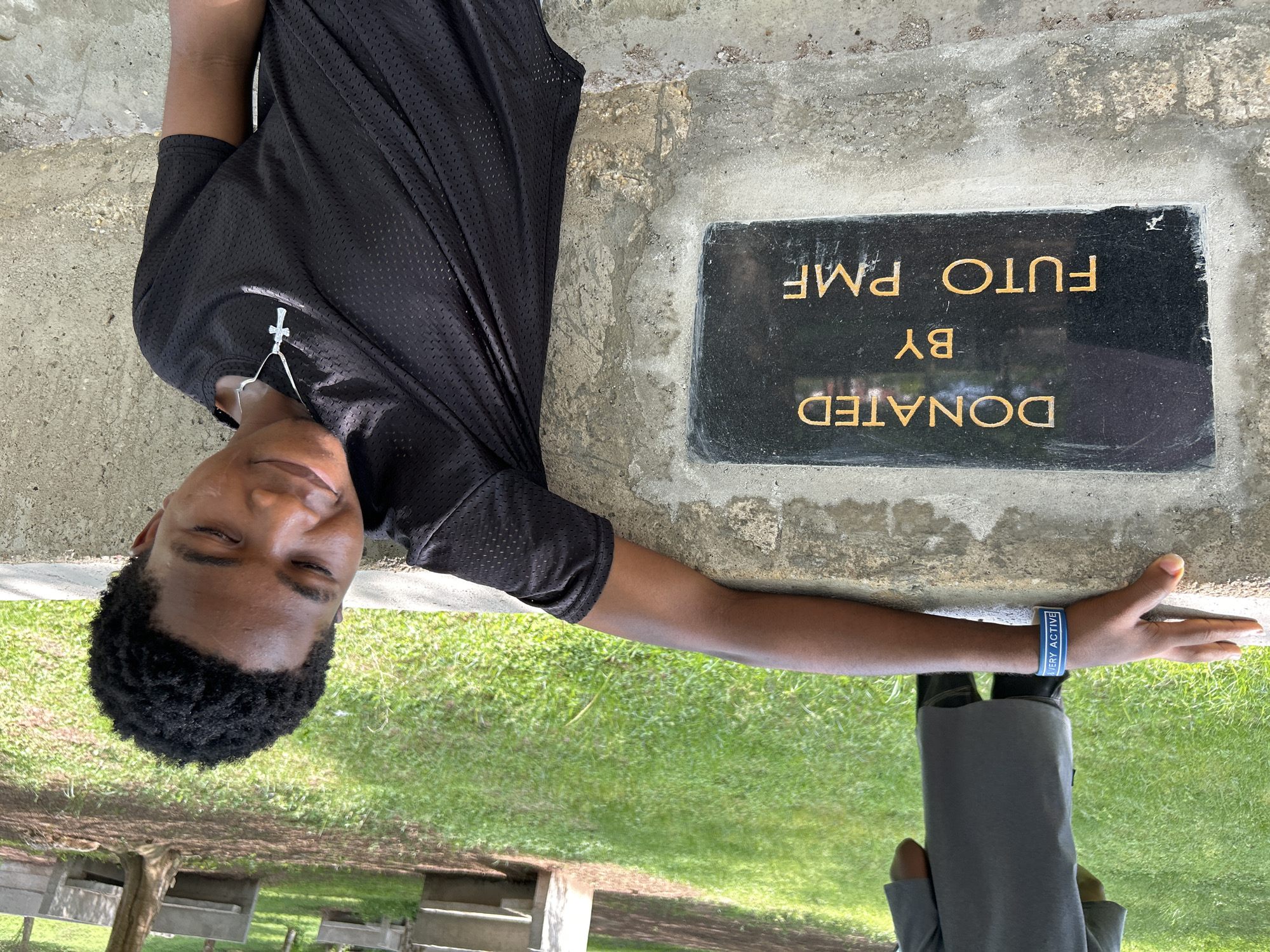 An outdoor scene features a young person, likely male, posing in bright daylight. The individual has dark skin and short, curly black hair. He is wearing a black mesh t-shirt and a silver cross pendant is visible on a chain around his neck. A blue and white wristband on his right wrist reads "VERY ACTIVE". He looks towards the camera with a relaxed smile, his eyes appearing closed or squinted, possibly due to the sun.
Beside him, embedded in a concrete surface, is a rectangular black plaque with gold lettering. The text on the plaque reads: "DONATED BY FUTO PMF". The surroundings consist of green grass and patches of bare earth. In the background, what appear to be concrete benches or structures are partially visible among trees, suggesting a park or public area. The lower body of another individual, dressed in dark trousers and light shoes, can be seen in the bottom right corner of the frame. The overall impression is a sunny daytime scene in Ihiagwa, Nigeria.