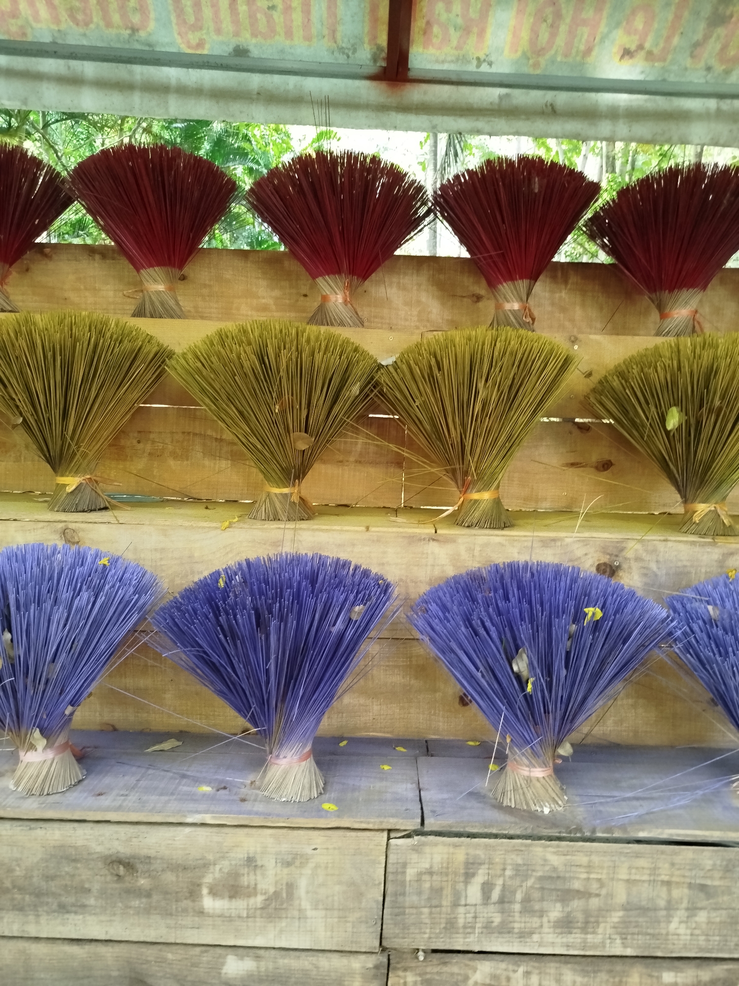 The image depicts bundles of incense sticks arranged on wooden shelves, likely for sale. Three tiers display red, yellow/green, and purple incense sticks, respectively. Each bundle is tied together with string. The scene is outdoors, evidenced by glimpses of trees in the background. Debris, such as leaves and small yellow petals, is scattered on the shelves and among the incense sticks. The photograph was taken in Tân Phú, Vietnam.