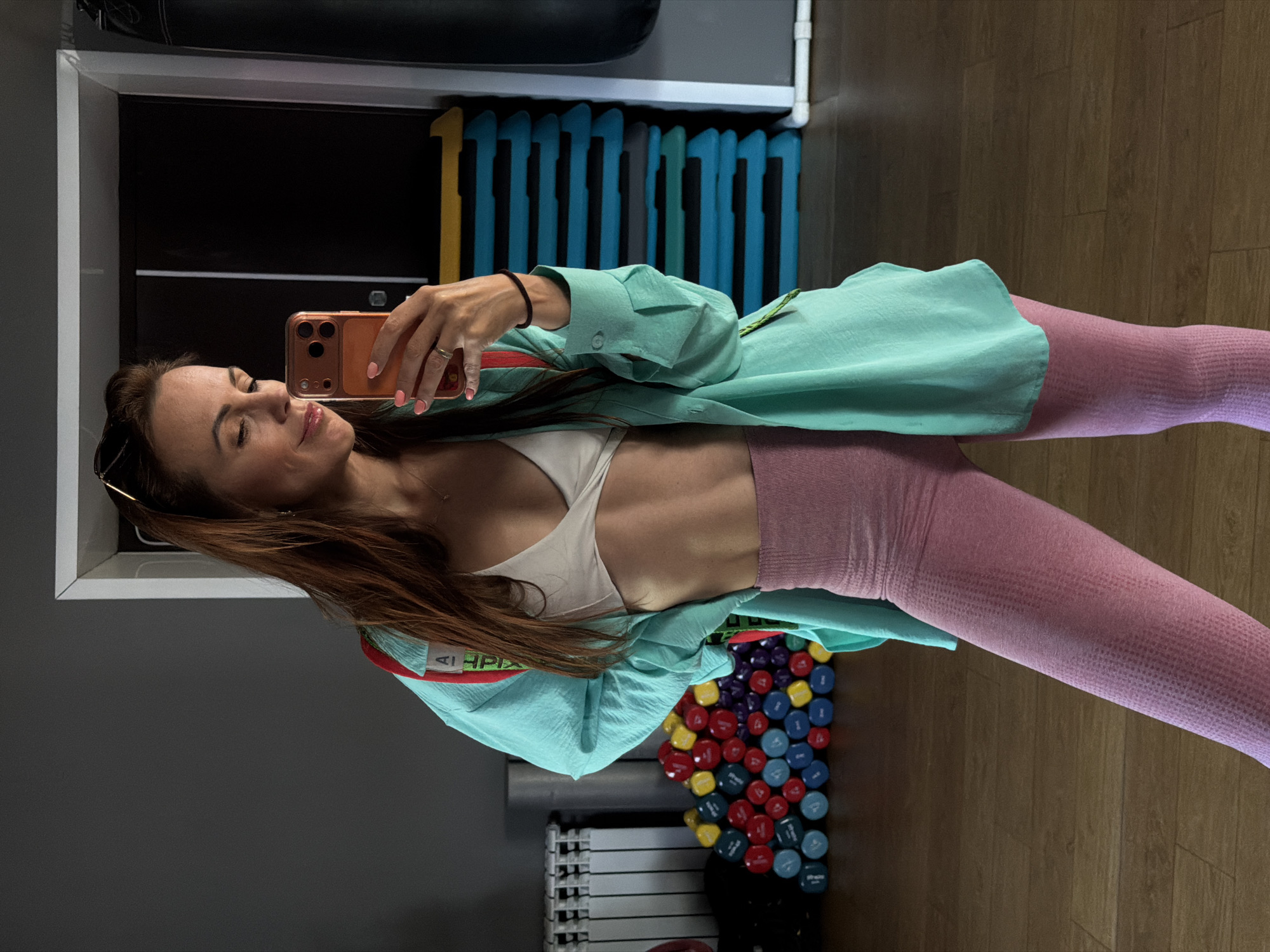 A woman stands in a gym, taking a mirror selfie with her phone. She is wearing a white sports bra, a teal oversized shirt, and pink leggings. In the background, there are weight plates, workout steps, and a punching bag. The gym has a grey wall and a wooden floor.