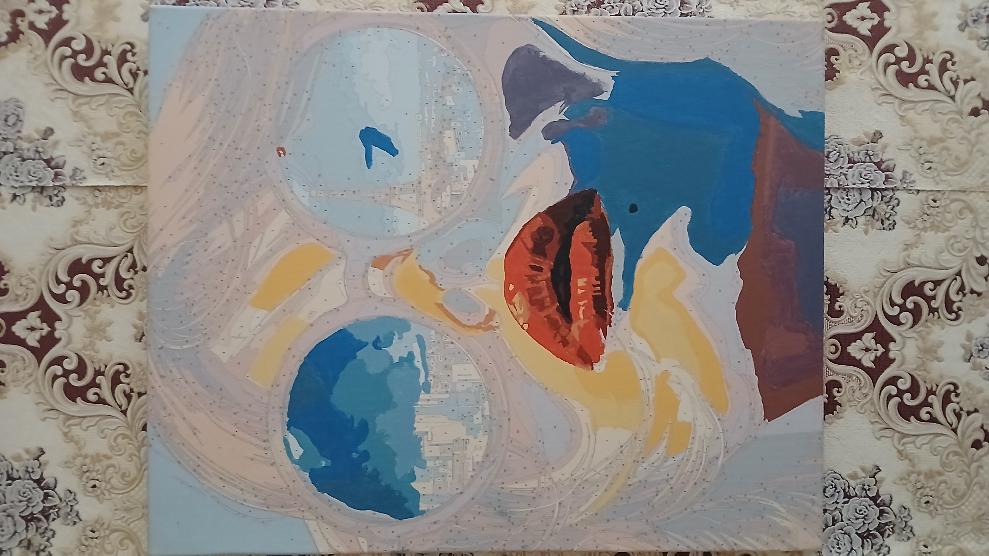 The image shows a partially completed paint-by-numbers canvas. The painting appears to be an abstract or surrealist artwork. It features three circular elements that resemble lenses or portals, with incomplete sections of blue and yellow colors. Above and to the right of these circles is a larger area filled with blue, grey, and brown paint. The background of the canvas is filled with small numbers corresponding to the paint-by-numbers guide. The canvas is placed on a surface covered with a patterned wallpaper or fabric.