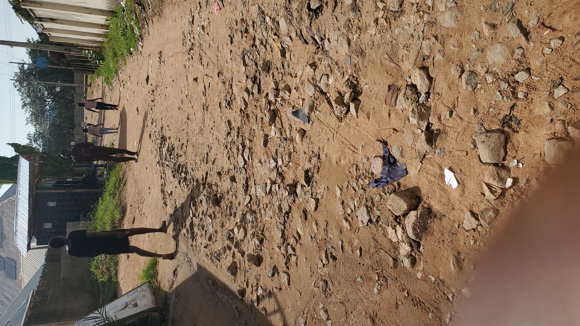 In Jalingo, Nigeria, a dirt road is depicted, characterized by loose sand and scattered rocks, indicative of uneven terrain. Several individuals are present, walking along the road. In the foreground, a person in a black dress walks away from the viewer, their posture suggesting movement. Further down the road, other figures are visible, contributing to the scene's depiction of daily life. Debris, including what appears to be dark fabric and white paper, is scattered on the ground, suggesting potential litter or discarded items. The environment includes buildings and vegetation bordering the road, with a clear sky above, indicating daytime.