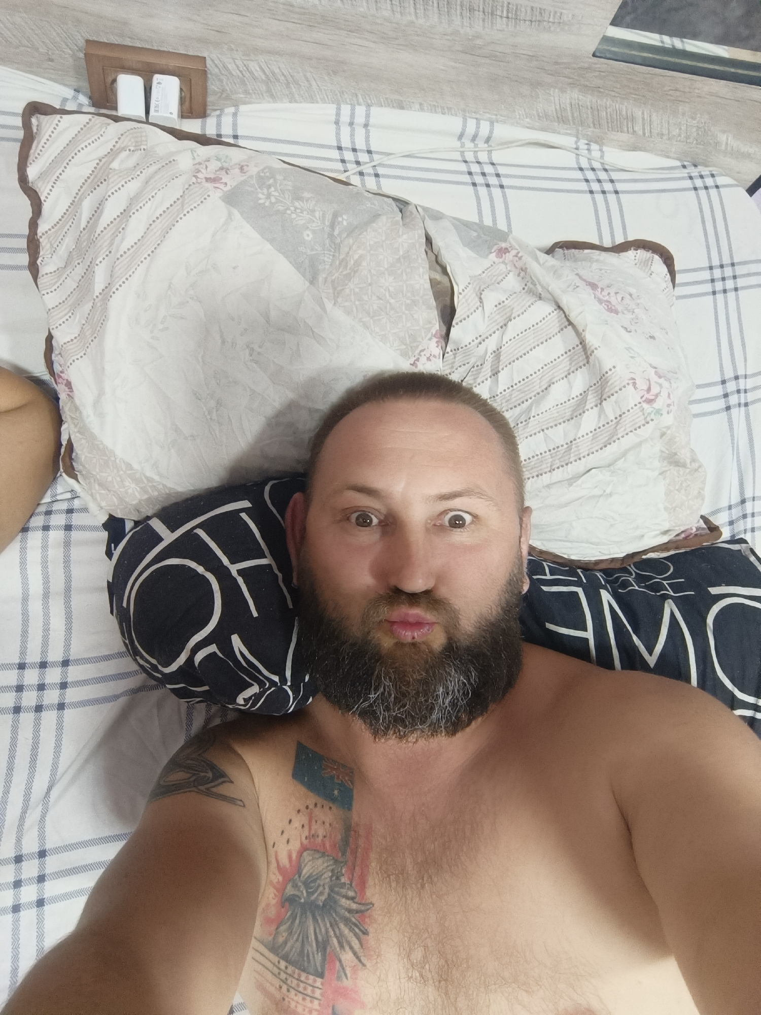 A man with a beard and a tattoo on his chest is lying in bed. He is looking at the camera with wide eyes and puckered lips. The bed is covered with a white and blue checkered comforter and a floral-patterned pillow. There are two white power adapters plugged into the wall behind the pillow. The room appears to be a bedroom. The time of day is not specified, nor is the weather. The location is not specified, but it is implied to be indoors. The man's expression suggests he is making a humorous or playful pose for the photo. There is no visible text in the image.