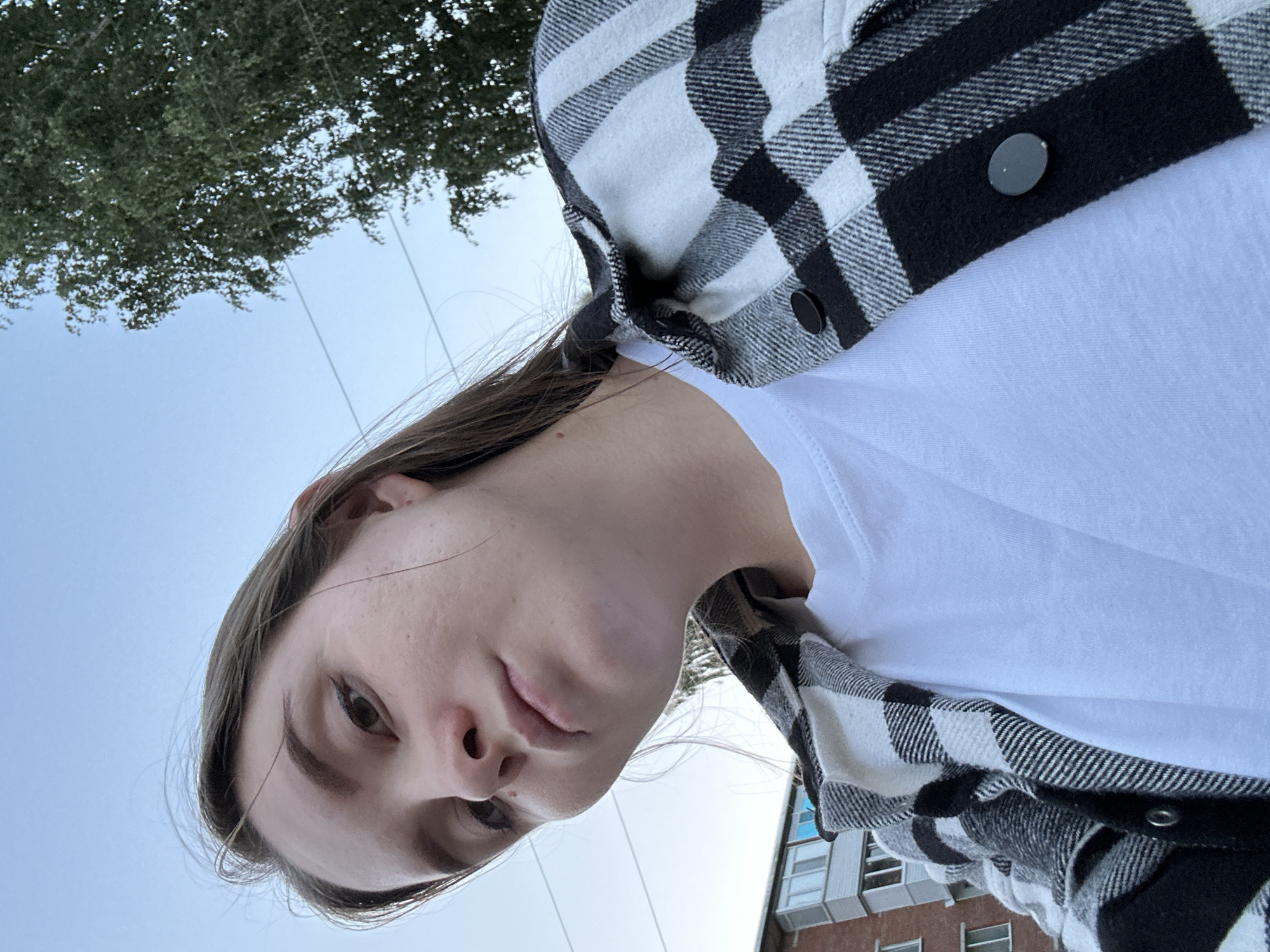 A low-angle selfie shows a young woman with long brown hair and light skin, wearing a white t-shirt and a black and white checkered flannel shirt. She is outdoors, with a building and trees visible in the background against a pale blue sky. The scene appears to be in Noviy, Russia, based on the architectural style of the building, which resembles typical residential blocks found in Russian cities. There is no indication of any specific action or interaction taking place; the image is a straightforward portrait.