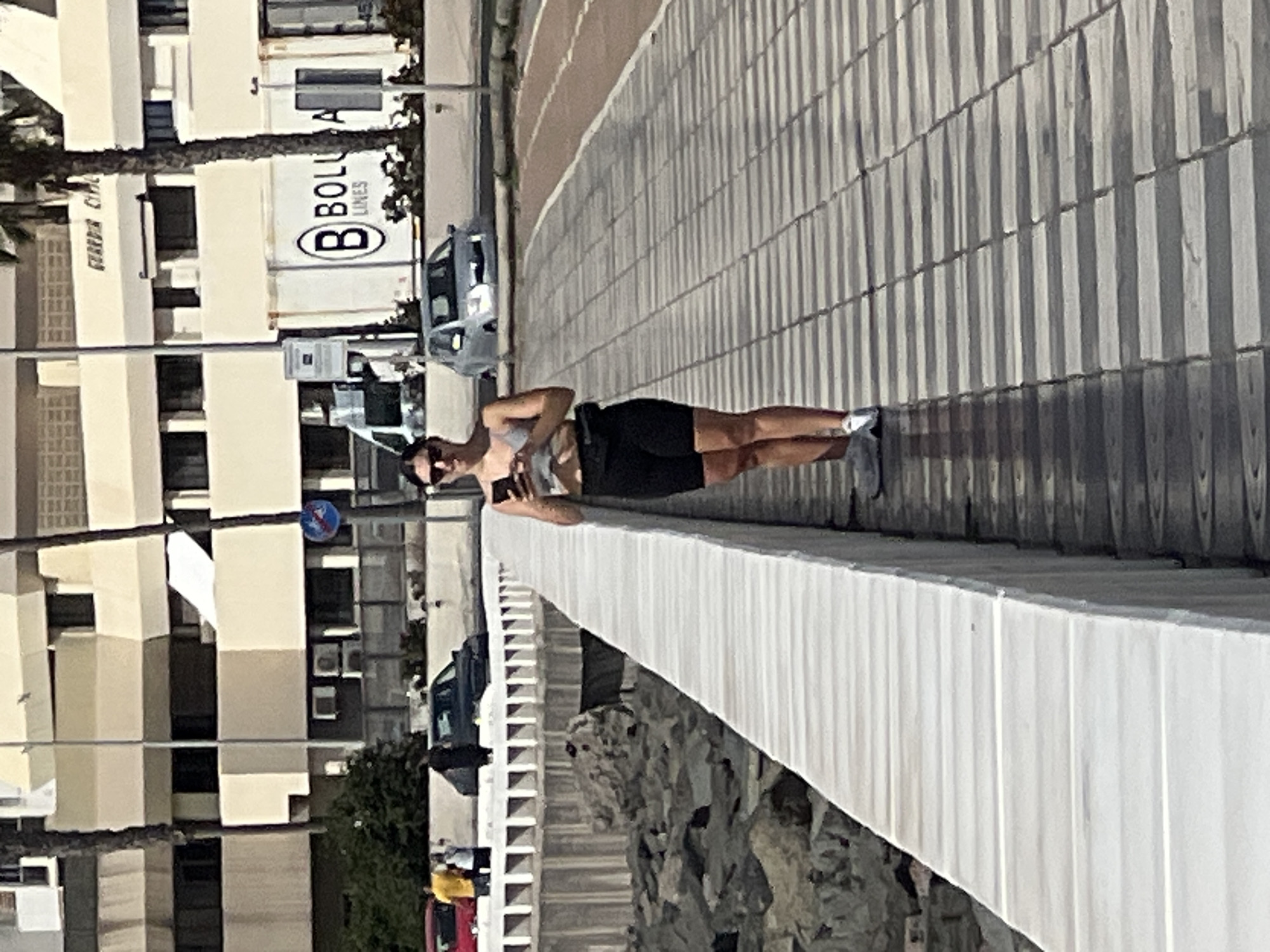 A woman stands on a seawall, holding a phone, in Las Palmas, Spain. She is wearing a gray top, black shorts, and white shoes. Behind her is a building with the name "BOLLORÉ LINES" on a container, as well as parked cars. The seawall is white, and the ground is tiled. The scene is outdoors, in daylight.