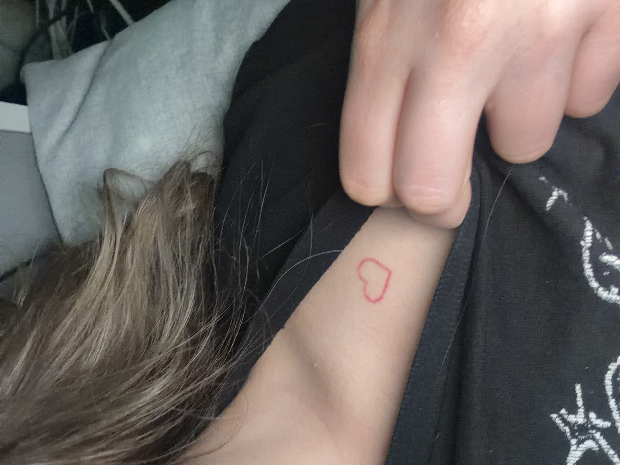 The image shows a person lying down. The person has blonde hair. They are wearing a black shirt. They have a small red heart tattoo on their upper left arm near their shoulder. Their fingers are visible near their shoulder, pulling up the shirt. A grey pillow or blanket is visible behind the person. The image does not provide information that would reveal the location of the city of Varna, Bulgaria.