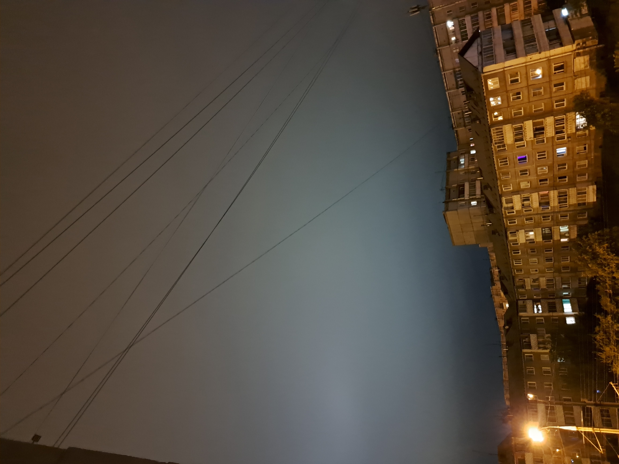 This is a nighttime, slightly angled, full-frame, eye-level shot of a city skyline in Irkutsk, Russia. In the foreground, a street lamp casts an orange glow on the left, illuminating a few trees and the lower portions of several residential buildings. The buildings are tall, multi-story structures with numerous windows, some of which are lit. They are a mix of concrete and panel construction, typical of Soviet-era housing.
In the mid-ground and background, more of these apartment blocks stretch across the frame, with the lights from their windows indicating occupancy. The sky above is dark and overcast, with a hazy, diffused light that obscures any stars or moon. Multiple power lines crisscross the upper portion of the image, silhouetted against the dim sky. The overall impression is of a dense urban environment at night, under a cloud-covered sky. There are no visible people in the image.
