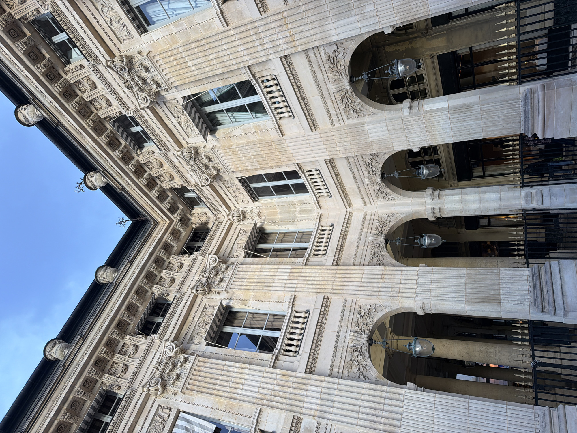 The image presents a low-angle shot of a Parisian building, likely a department store or a high-end commercial structure, evidenced by its ornate architectural details. The facade features intricate stonework, including decorative relief patterns, columns, arched openings, and balustrades. Windows punctuate the facade, exhibiting a mix of reflective surfaces. The building is light-colored, suggesting limestone or a similar material typical of Parisian architecture. Hanging light fixtures are visible within the arched openings. The sky is blue and clear, contrasting with the building's color. The image captures the grandeur and detail of Parisian architecture. The scene is devoid of people.