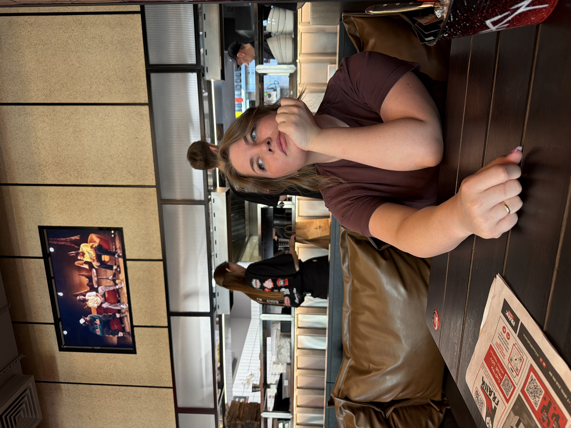 The image depicts an indoor scene at a "Frank By Basta" restaurant in Kazan, Russia, identifiable by Cyrillic text and the "+7 (843)" area code on a menu. In the foreground, a young woman with light brown hair, freckles, and blue eyes is seated at a dark brown slatted wooden table, wearing a dark maroon t-shirt. Her chin rests on her left hand, displaying a gold ring on her left ring finger and light pink nail polish on her left index finger. She gazes upwards and to the left with a contemplative expression. A folded paper menu, printed with "FRANK" branding and QR codes, lies on the table to her left, alongside a small red circular object and a red speckled bucket holding cutlery on her right.
Behind the woman, a brown leather-upholstered booth or bench seat is visible. The background reveals an open kitchen or service counter area. Three individuals, presumably staff, are present behind the counter. To the left, a woman with long dark hair, wearing a black t-shirt with multiple colorful patches and a black apron, stands facing away from the camera, appearing to handle a brown paper bag. In the center, a man with short brown hair is seen from the back. To the right, another man wearing a black baseball cap is visible in profile.
Above the counter, the wall features light brown textured panels with dark vertical dividing lines. A framed television screen is mounted on this wall, displaying an image or video of three men seated on red chairs or sofas in a staged interior setting. Below the main wall, horizontally ribbed or fluted translucent panels are visible, separating the upper wall from the service area which includes stainless steel surfaces, shelves with kitchenware, and overhead lighting fixtures. A square air vent is visible in the ceiling in the upper left portion of the image.