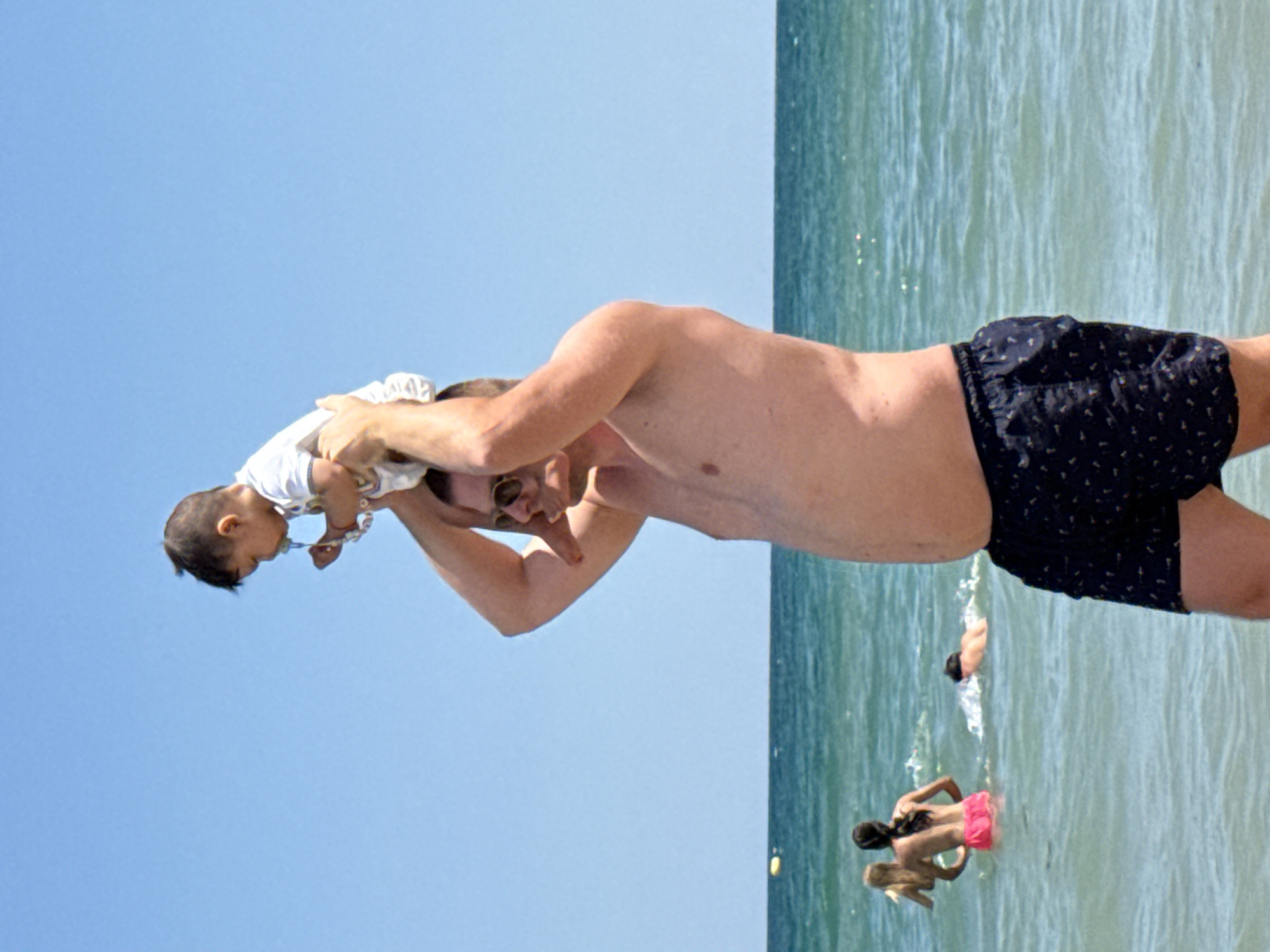 A man is standing on a beach in Kranevo, Bulgaria, holding a baby on his shoulders. The man is wearing dark patterned swim trunks and sunglasses. The baby is wearing a white onesie and has a pacifier. The ocean is calm and blue, with a few other people swimming in the background. The sky is clear and bright blue. The scene appears to be a sunny day at the beach.
