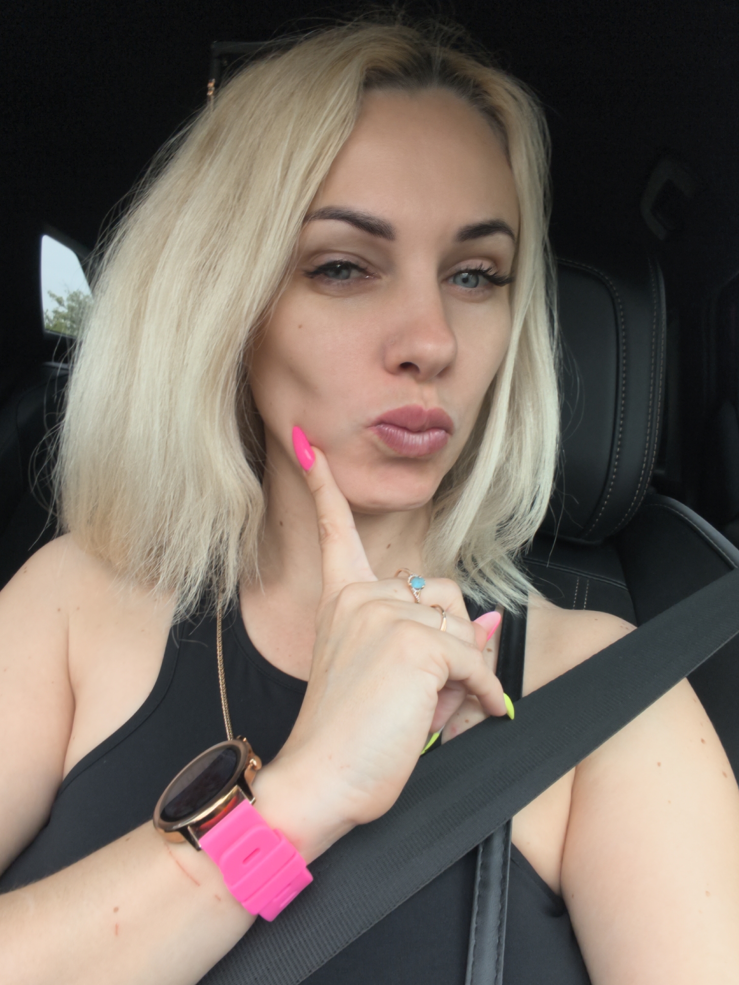 A woman with light blonde hair, wearing a black tank top and a seatbelt, poses in what appears to be a car interior. Her expression is a slight pout, and she touches her cheek with a finger adorned with pink nail polish. She also wears a gold-colored wristwatch with a pink band and multiple rings on her other hand, one with a turquoise stone. The environment suggests the interior of a vehicle, with black leather seats and a view through the window showing foliage. There is no specific information in the image to indicate a location in Novorossiysk, Russia.
