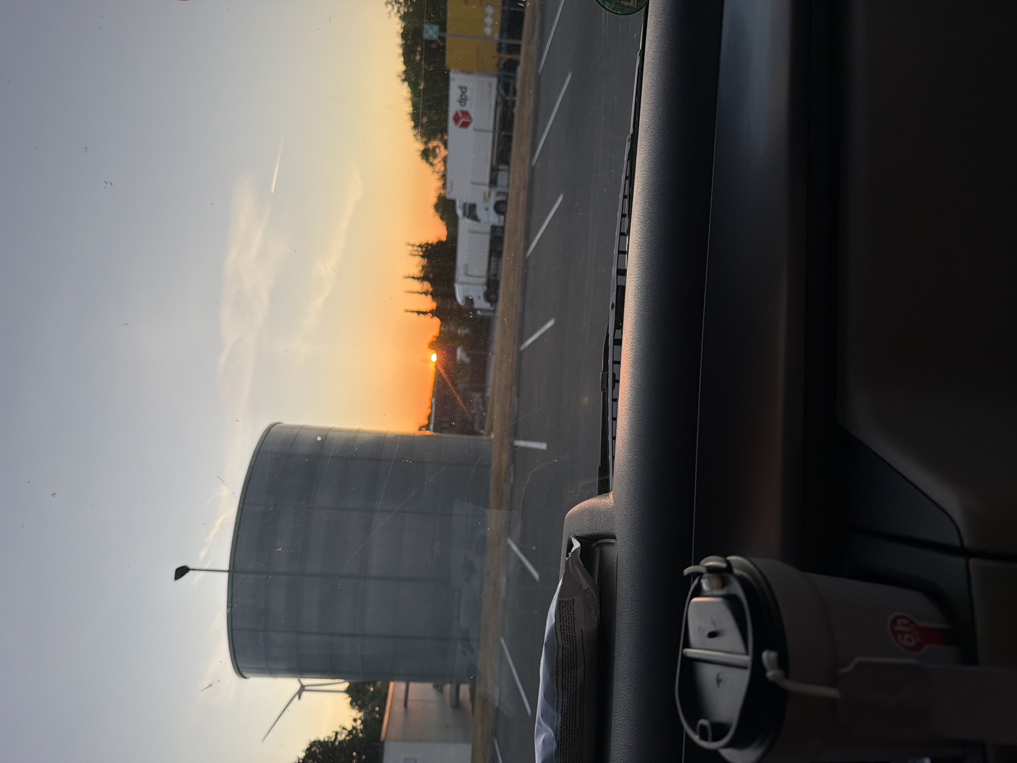 The image depicts a sunset view through a car's windshield. In the foreground, the dashboard is visible, with a travel mug and a packet of tissues resting on it. Beyond the car, a large, cylindrical silo-like structure stands prominently. To the left, a wind turbine is partially visible. In the background, several white trucks are parked in a lot, one of which displays the "dpd" logo. The sky is a gradient of orange and blue, indicative of sunset. The scene appears to be set in an industrial or commercial area, possibly near a logistics hub or storage facility. The location is identified as Witten, Germany.