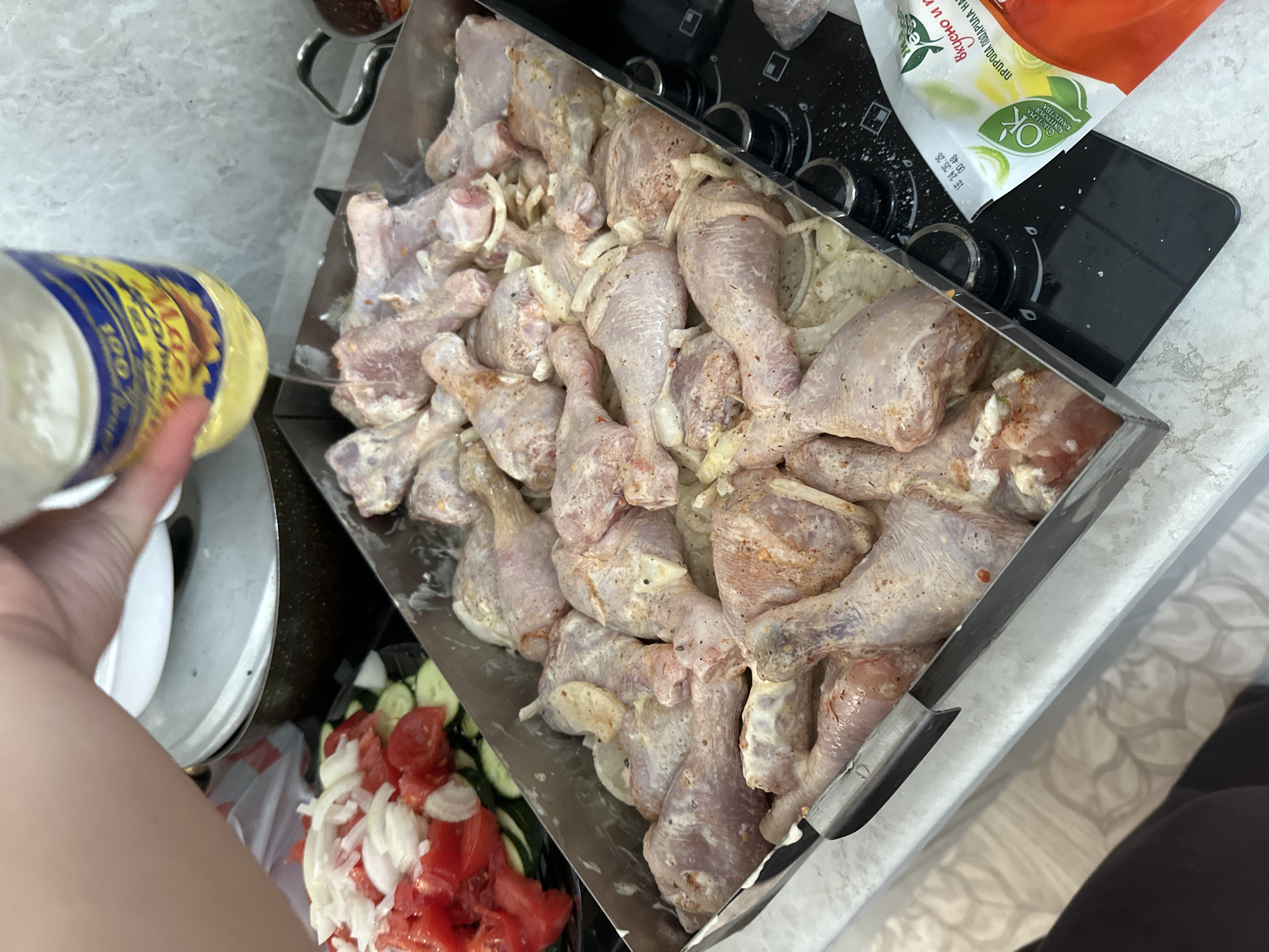 In the city of Voznesenka, Russia, a close-up shot captures the preparation of chicken legs. The raw chicken legs, coated in a creamy marinade, are arranged in a large metal baking pan. Sliced onions are visible underneath the chicken. To the left, a salad of chopped tomatoes and cucumbers is prepared in a clear plastic container. A hand is in the process of drizzling oil from a bottle onto the chicken. The scene takes place on a kitchen countertop, with a stovetop visible in the background, suggesting a home kitchen environment.