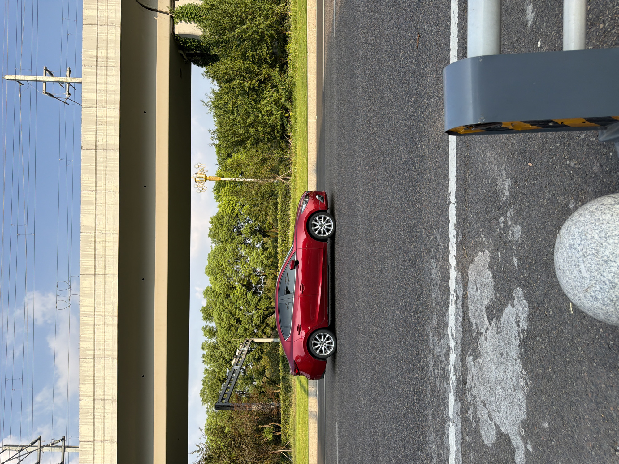 A red sedan is driving on a road under a large overpass. The car is in motion, viewed from the side. The road is asphalt with a white line. The overpass is made of concrete. Trees and greenery line the side of the road beyond the overpass. In the background, power lines and poles are visible against a blue sky with clouds. A street lamp with multiple globes is also present near the trees. In the foreground, a gray bollard with yellow and black markings is visible on the right side of the image. The scene appears to be an urban or suburban environment in Jiangjin, China, given the architectural style of the overpass and the surrounding vegetation.