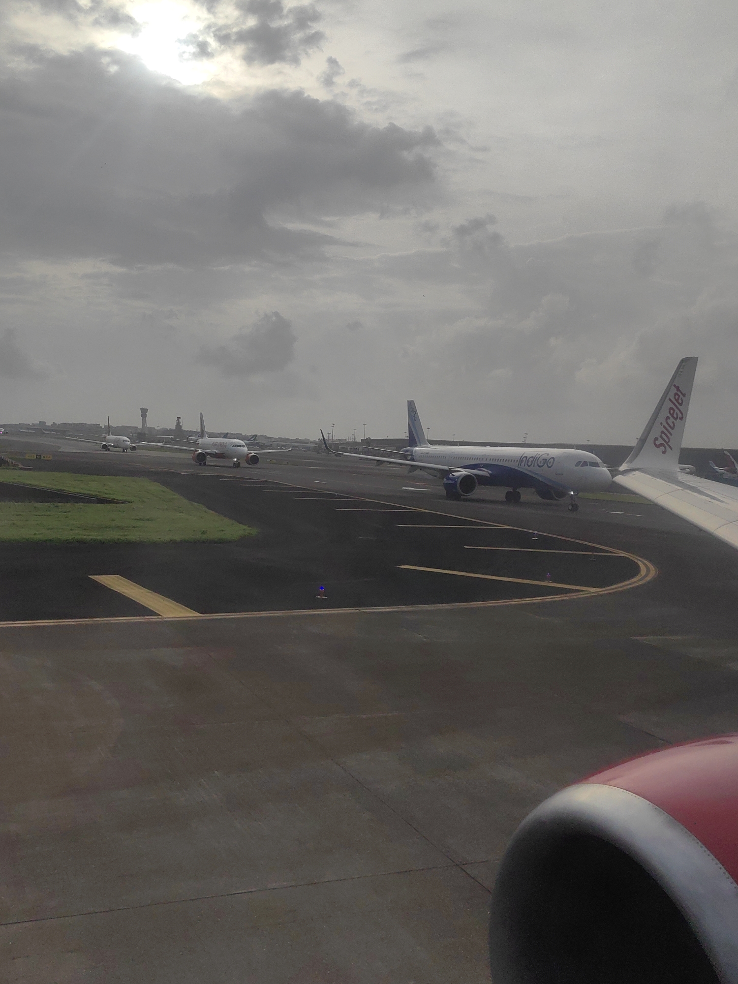 The image depicts a tarmac scene at Chhatrapati Shivaji Maharaj International Airport in Mumbai, India, under an overcast sky with the sun peeking through the clouds. Several airplanes are visible on the runway and taxiways. In the foreground, the red engine cowling of an aircraft is partially visible on the right side. Further down the tarmac, a white airplane with "Indigo" branding on its fuselage is positioned centrally. To the left of the Indigo plane, another aircraft, possibly a SpiceJet plane, is seen with its tail fin labeled "SpiceJet." In the distance, more aircraft are lined up, and airport infrastructure, including control towers, is discernible against the cloudy sky. The overall scene suggests an active airport environment, possibly during a pause in operations or before takeoff/after landing.