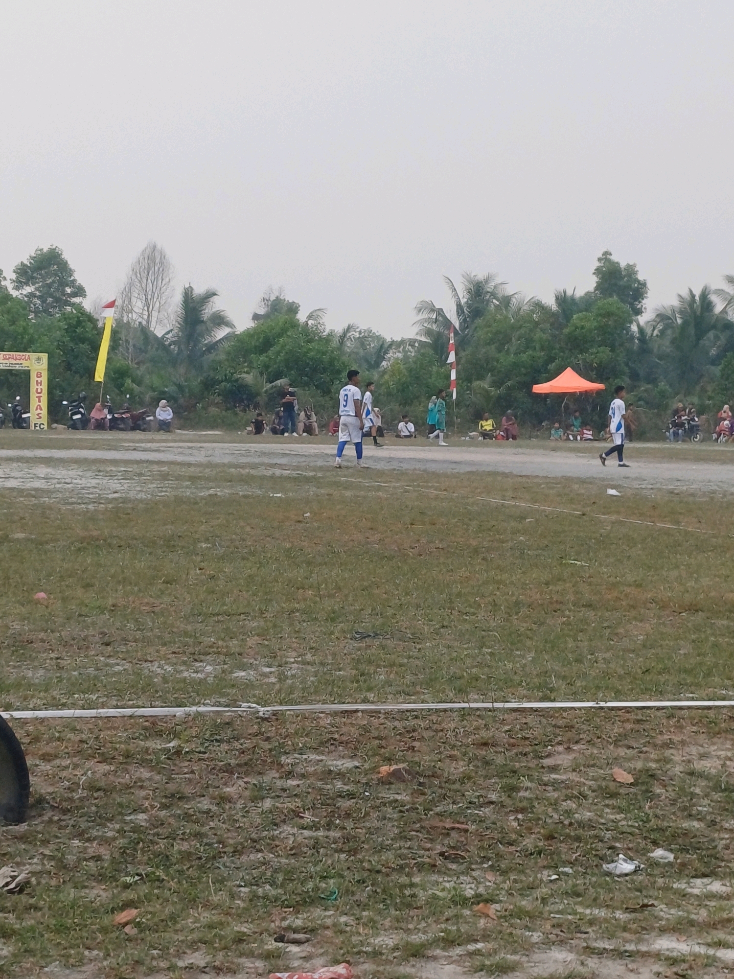 The image captures a scene at a football field in Dumai City, Indonesia, under an overcast sky. In the foreground, a grassy field with patches of dirt is visible, bordered by a white plastic barrier. Several individuals, some in football attire, are present on the field and along the sidelines. To the left, a banner with text is partially obscured by trees and flags, including a yellow and red striped flag. Various motorcycles are parked behind the banner, and a few spectators are seated or standing in the background. An orange tent is set up further back, providing shade. The overall environment suggests a casual sporting event taking place in an outdoor, somewhat rustic setting.