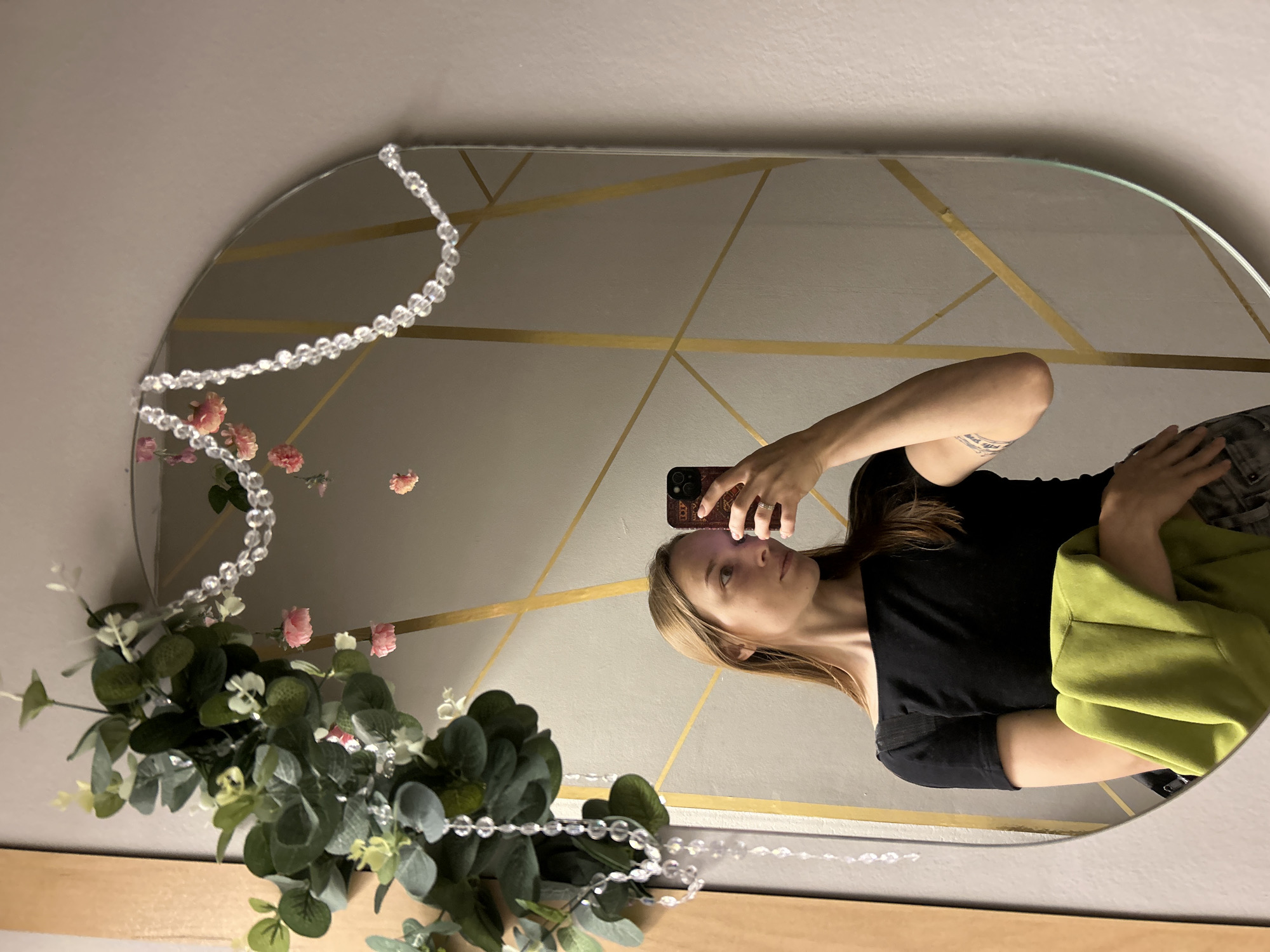 A young woman with long blonde hair is taking a selfie in an oval mirror. She is wearing a black off-the-shoulder top and holding a lime green garment over her arm. Her left hand is resting on her hip. The mirror has a decorative garland of artificial eucalyptus leaves and small pink flowers draped across the top, with a strand of clear beads hanging down. The wall behind the mirror has a geometric pattern of gold lines. The woman is positioned in front of a wooden doorframe. The scene is indoors, likely in a residential setting in Chaikovsky, Russia.